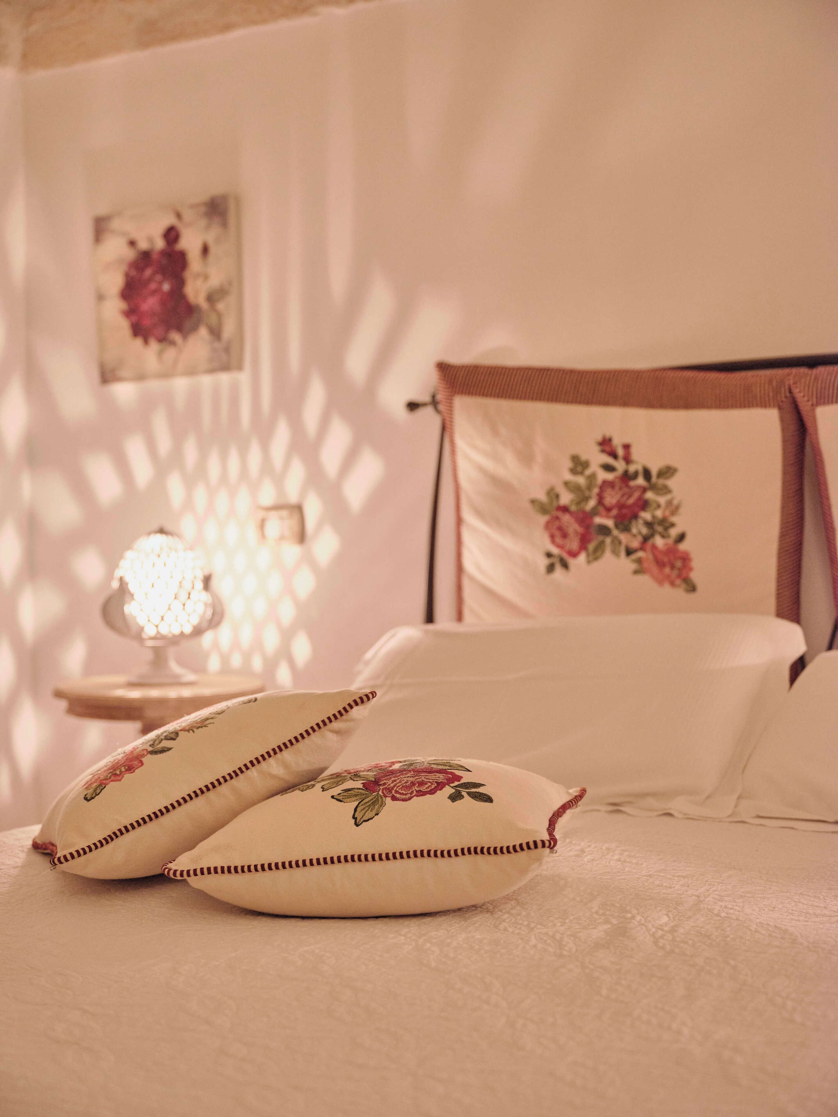 A bedroom with a bed, pillows, a nightstand, a lamp, and a wall with a floral painting.