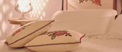 A bedroom with a bed, pillows, a nightstand, a lamp, and a wall with a floral painting.