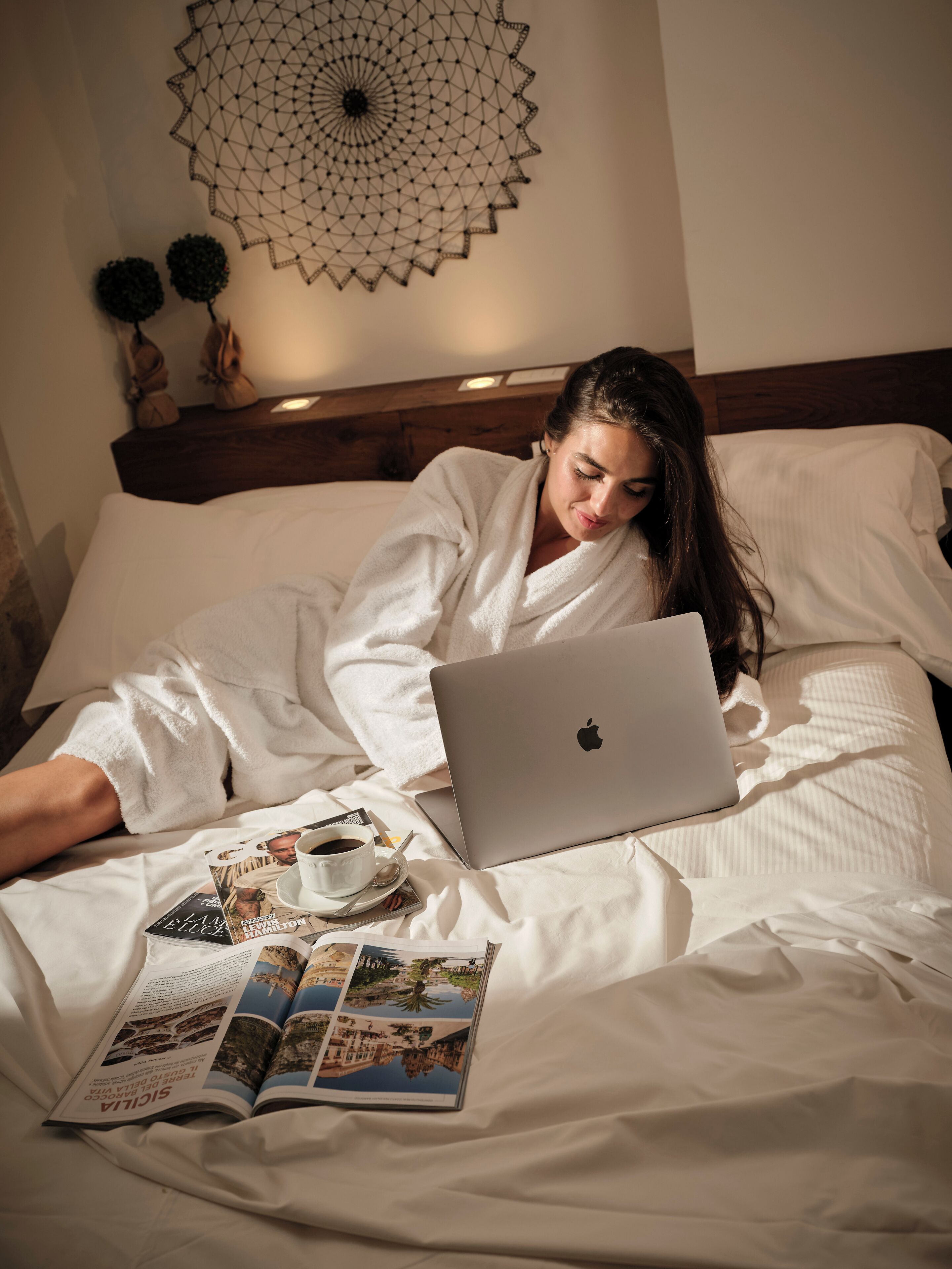 A person lying on a bed with a laptop, a cup of coffee, and a magazine.