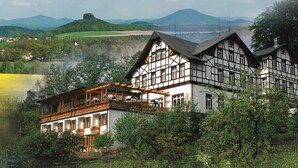 A traditional half-timbered building with a balcony overlooking a lake, surrounded by lush greenery and hills in the background. - Panoramahotel Wolfsberg (Reinhardtsdorf-Schoena)