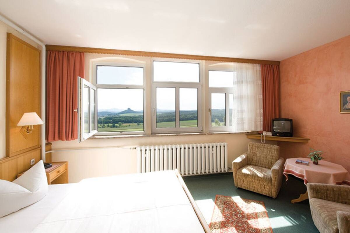 A hotel room with a bed, two armchairs, a small table, a TV, and a window with a view of the countryside.