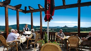 A terrace with wicker chairs and tables, a flag, and a view of the countryside. - Panoramahotel Wolfsberg (Reinhardtsdorf-Schoena)