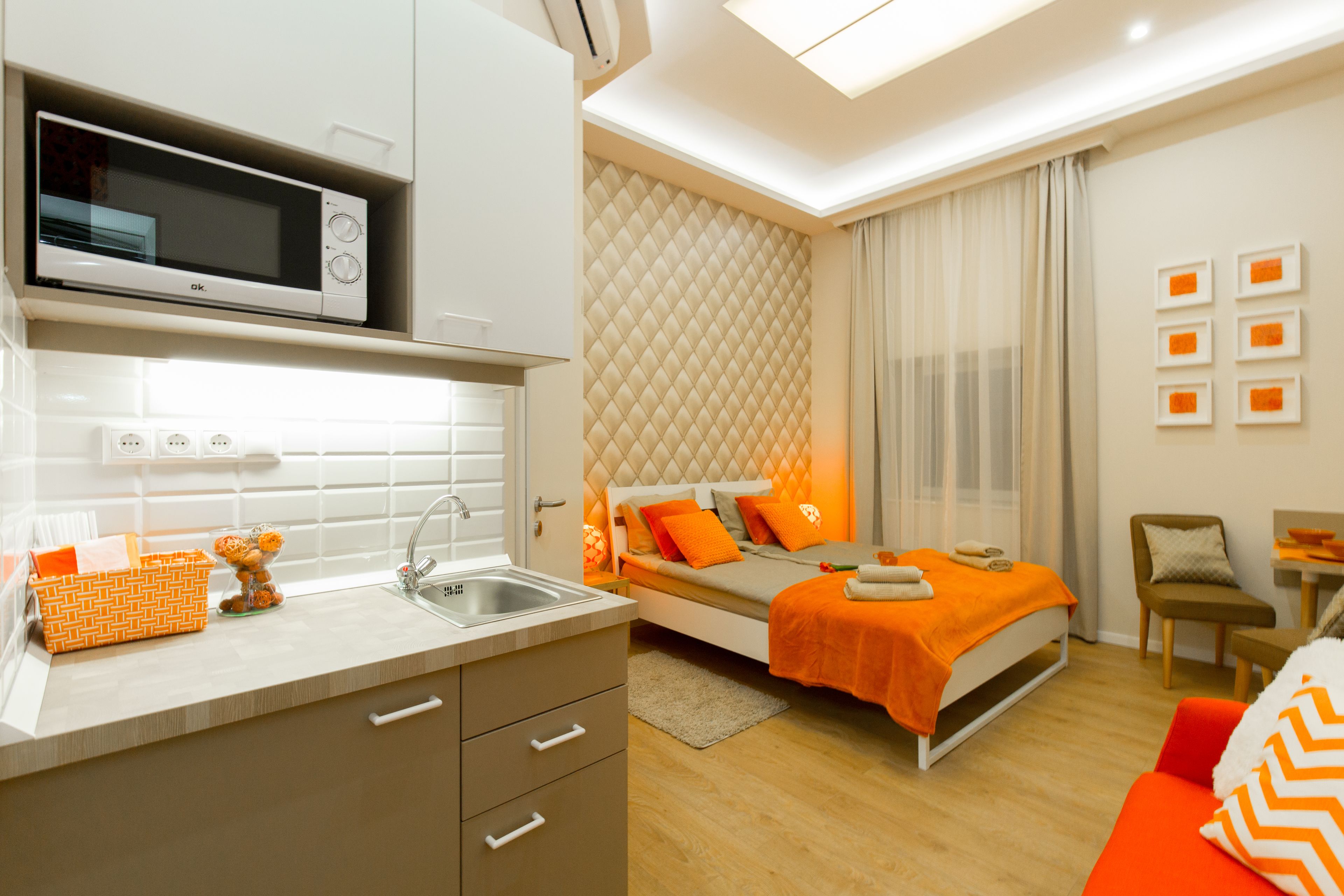 A compact hotel room with a kitchenette, a bed with orange bedding, a small seating area, and a view of the cityscape.