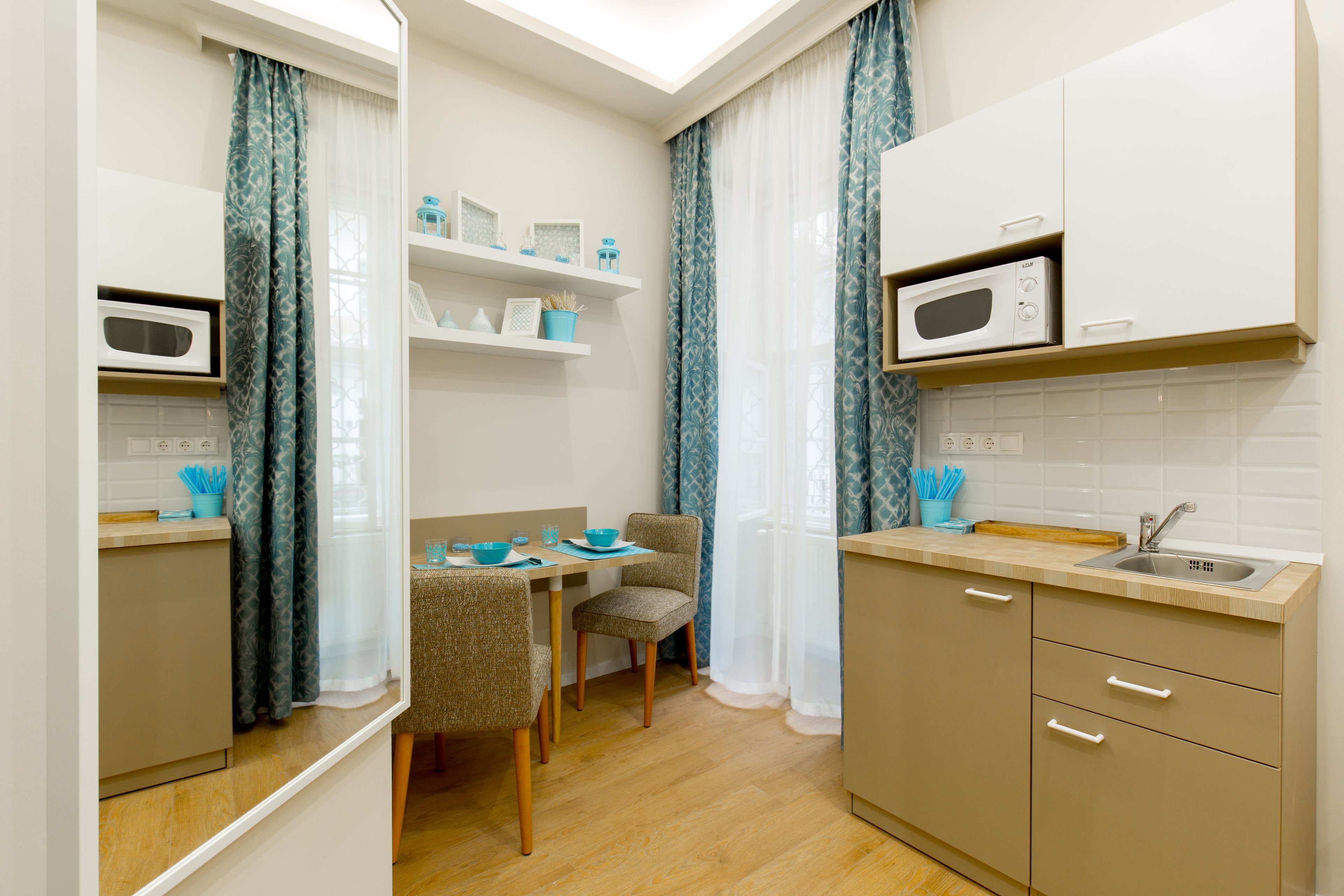 Cocina compacta con microondas, pileta y muebles de madera. Se aprecia una mesa de comedor con sillas y una ventana con cortinas.
