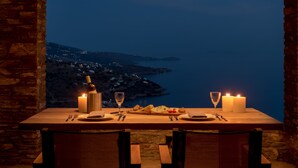 A table set for two with candles, a bottle, and a glass, overlooking a coastal view. - Andros Tessera (Andros)