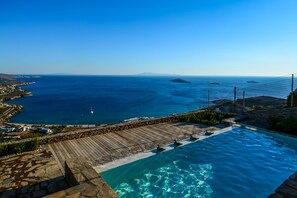 Una piscina con agua azul cristalina, rodeada de muros de piedra y una terraza de madera, con vistas a una zona costera con edificios y el mar.