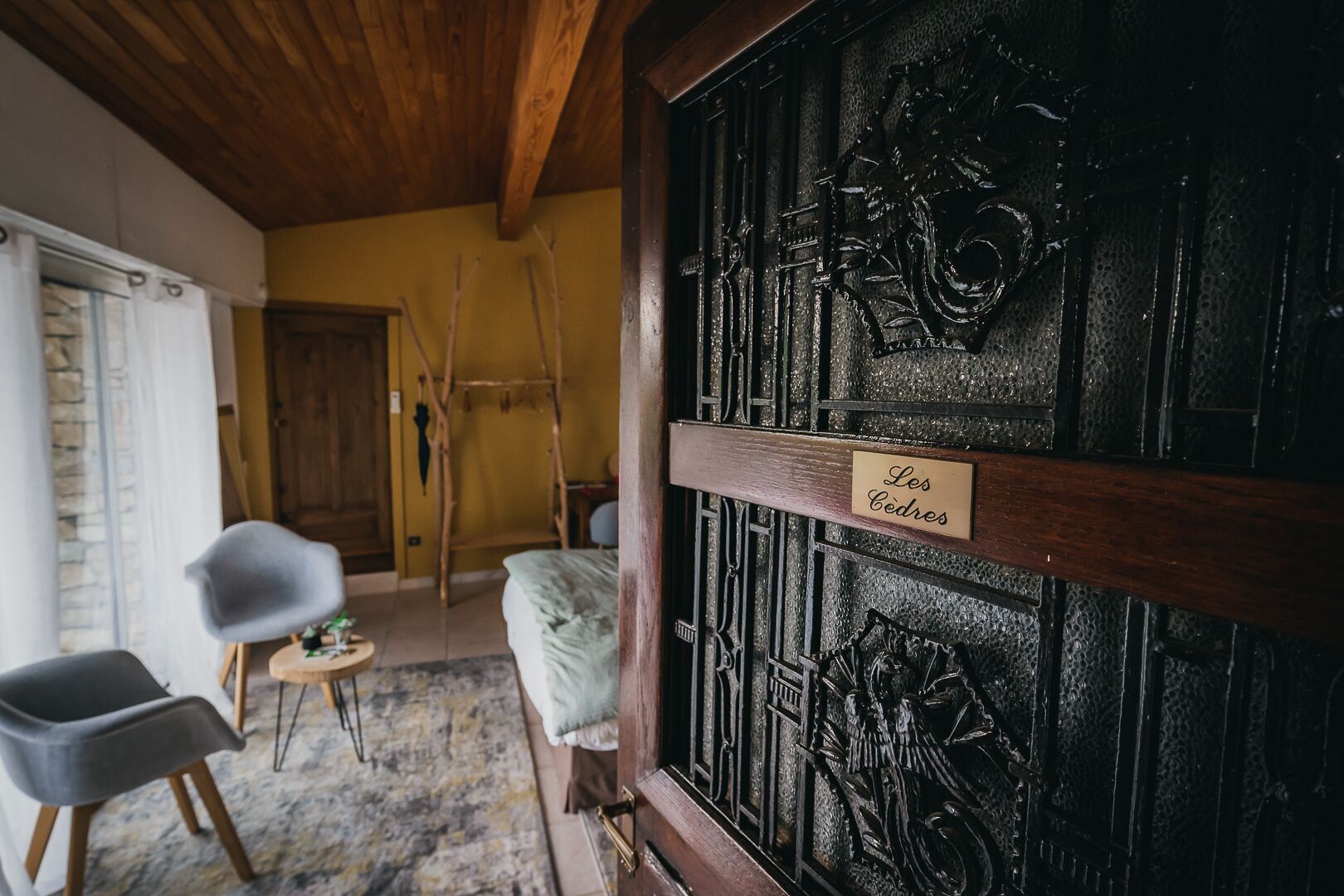 Ein Zimmer mit Holzbalkendecke, einem Bett mit weißer Decke, einem kleinen runden Tisch mit einem Stuhl und einer Tür mit dem Schild "Les Cendres".