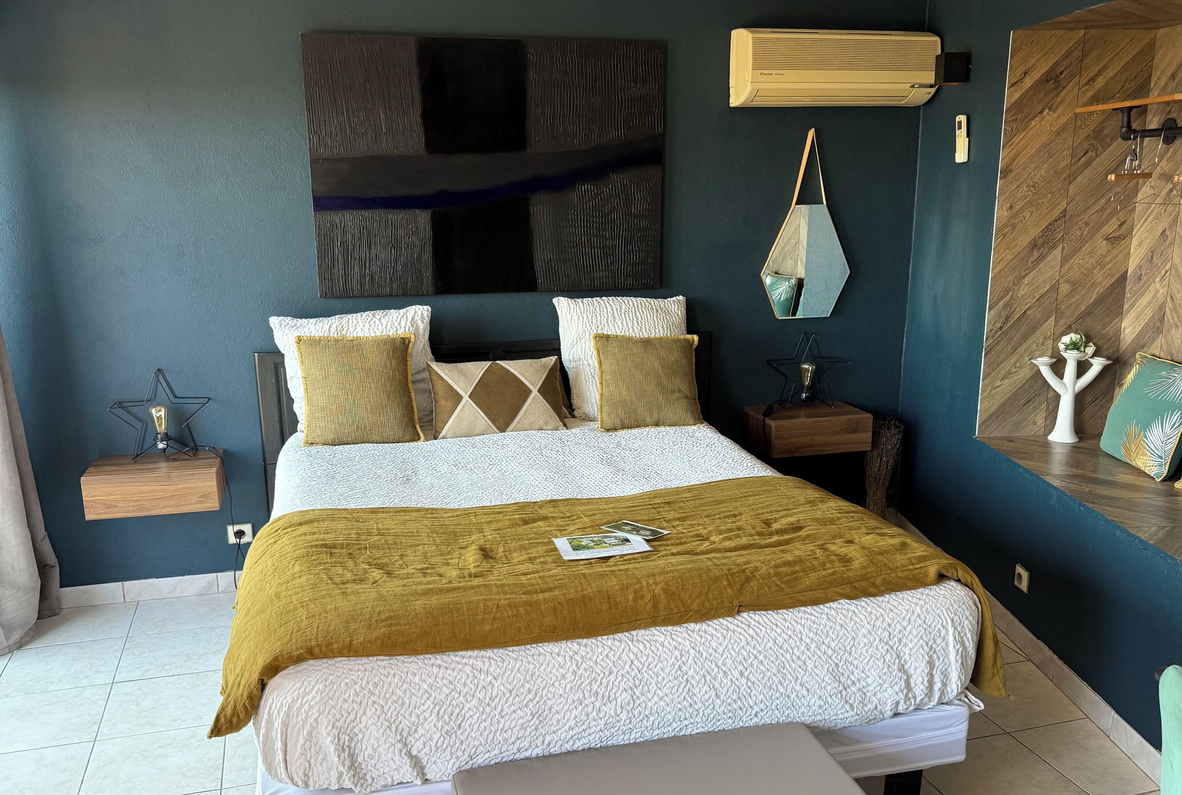 A neatly made bed with a mustard-colored throw blanket, a large abstract painting above it, and a wooden nightstand with a star-shaped lamp beside it.