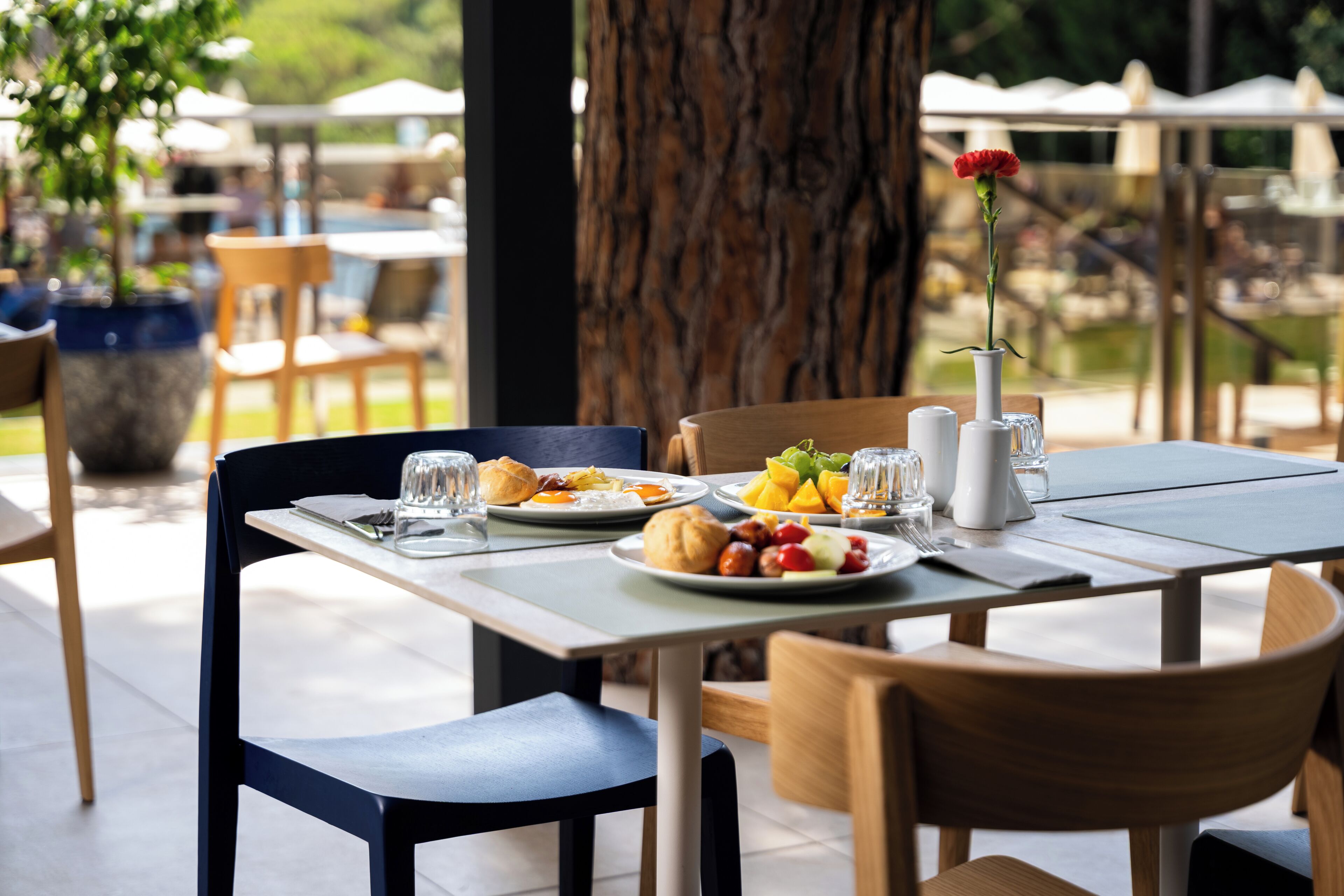 An outdoor dining area with a table set for breakfast, including plates of food, glasses, and a vase with a flower.