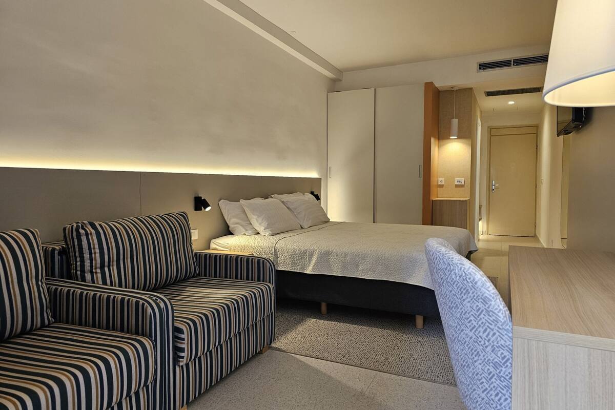 A modern hotel room with a striped sofa, a bed with a white duvet, and a wooden wardrobe.