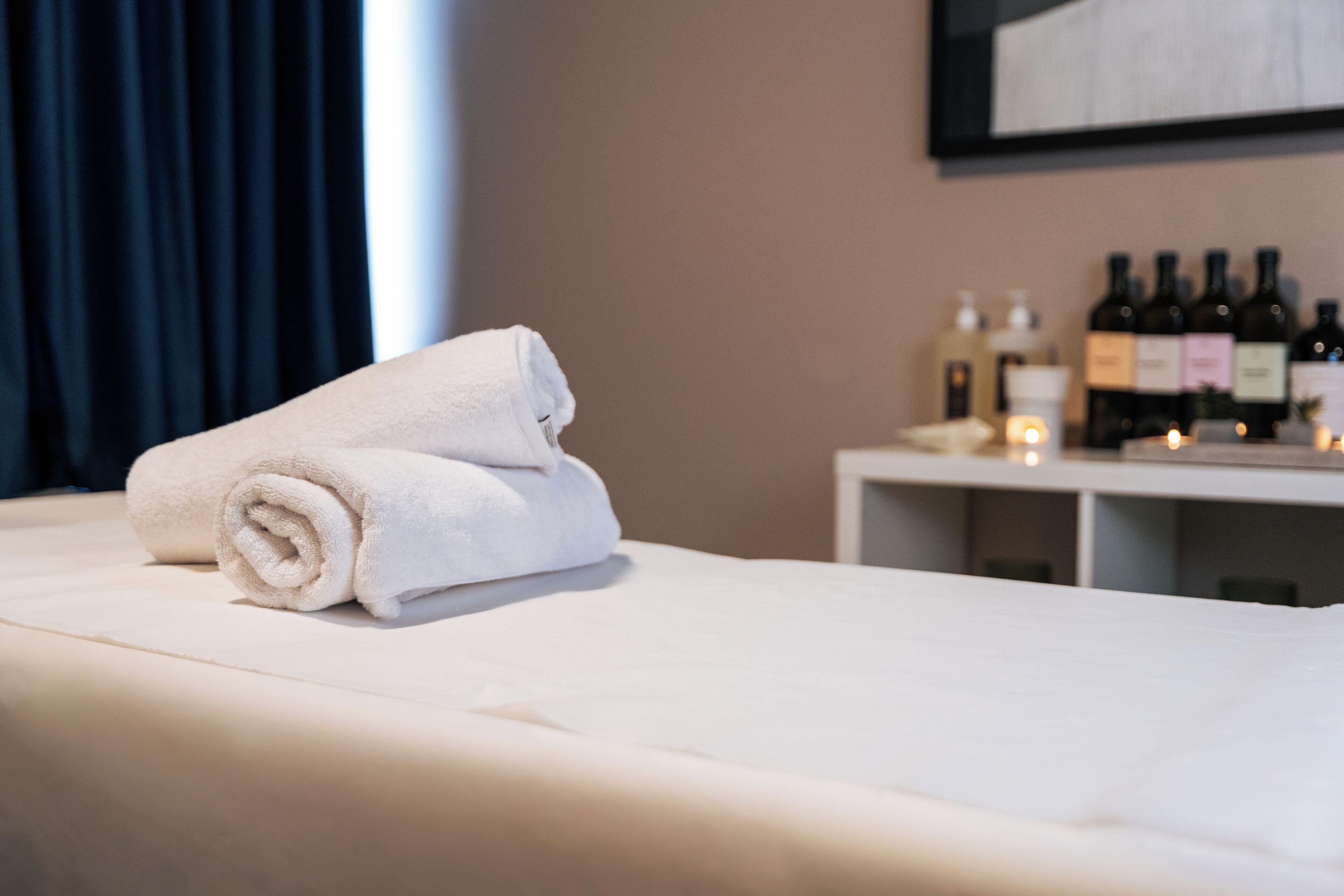 A neatly made bed with a rolled white towel, a bedside table with bottles, and a wall-mounted shelf with more bottles.