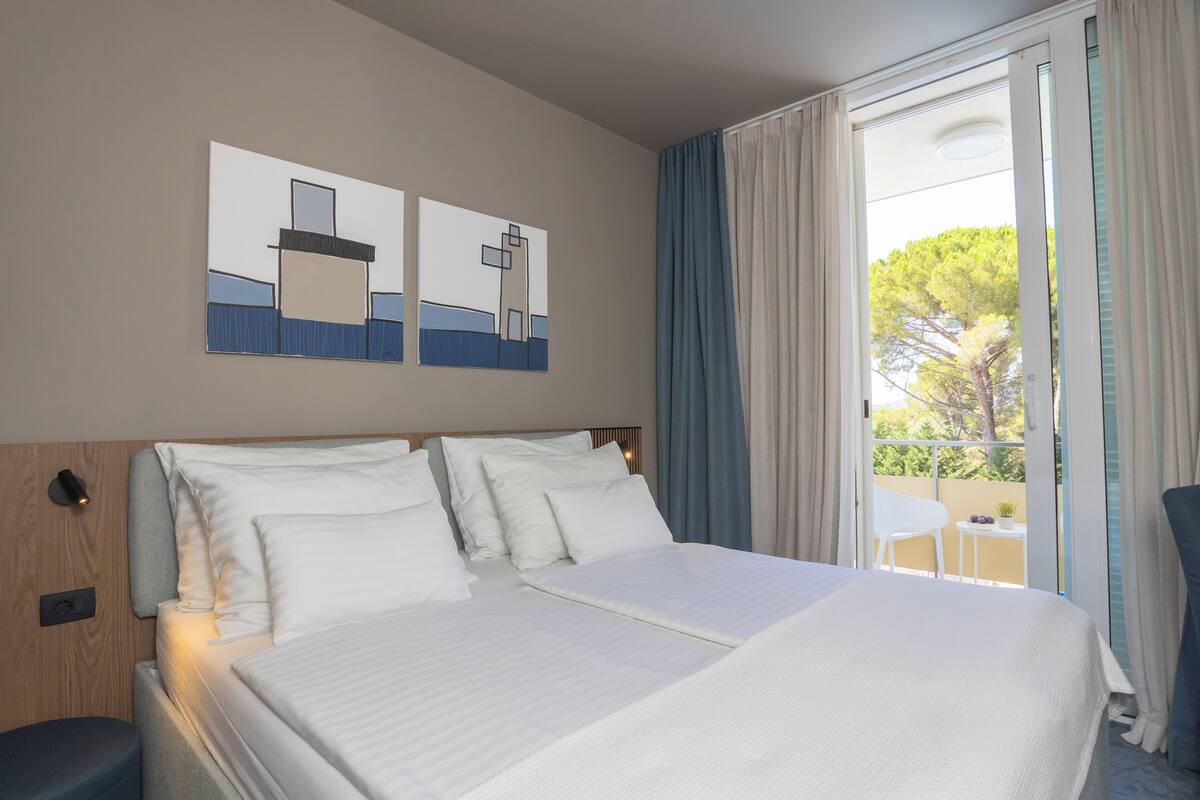 A hotel room with a large bed, a wooden headboard, and two wall art pieces. There is a balcony with a table and chair, and a view of trees outside.