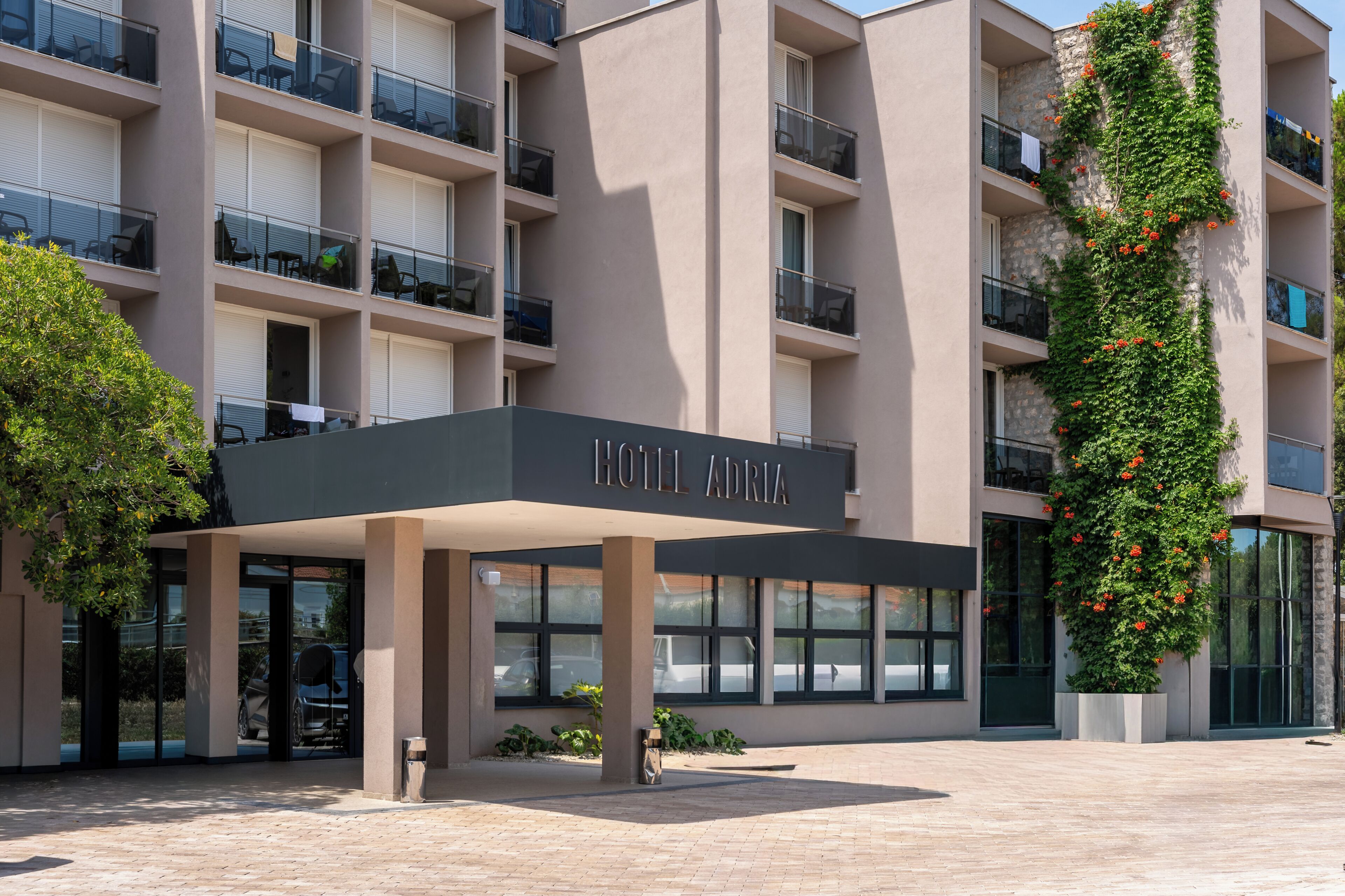 A hotel entrance with a covered area, multiple balconies, and a sign reading 'HOTEL ADRIA'.