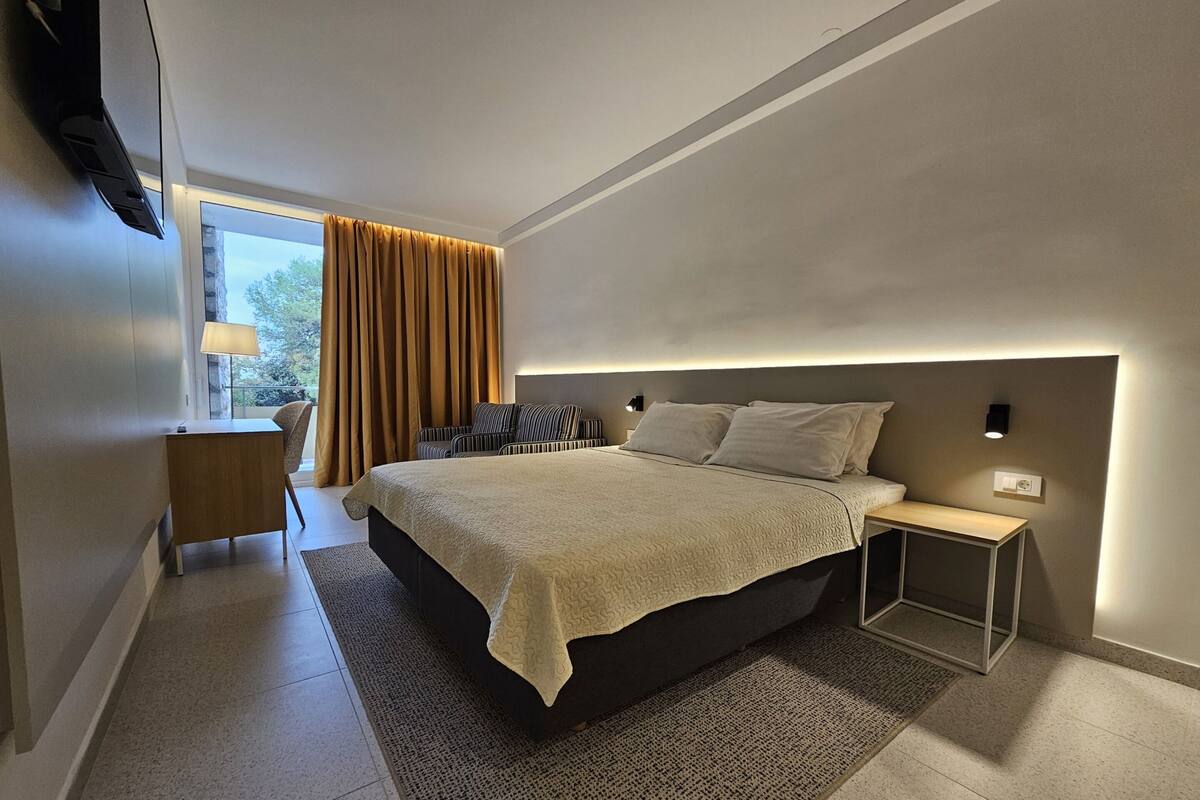 A modern hotel room with a large bed, a bedside table, a desk with a chair, and a view of the outdoors.