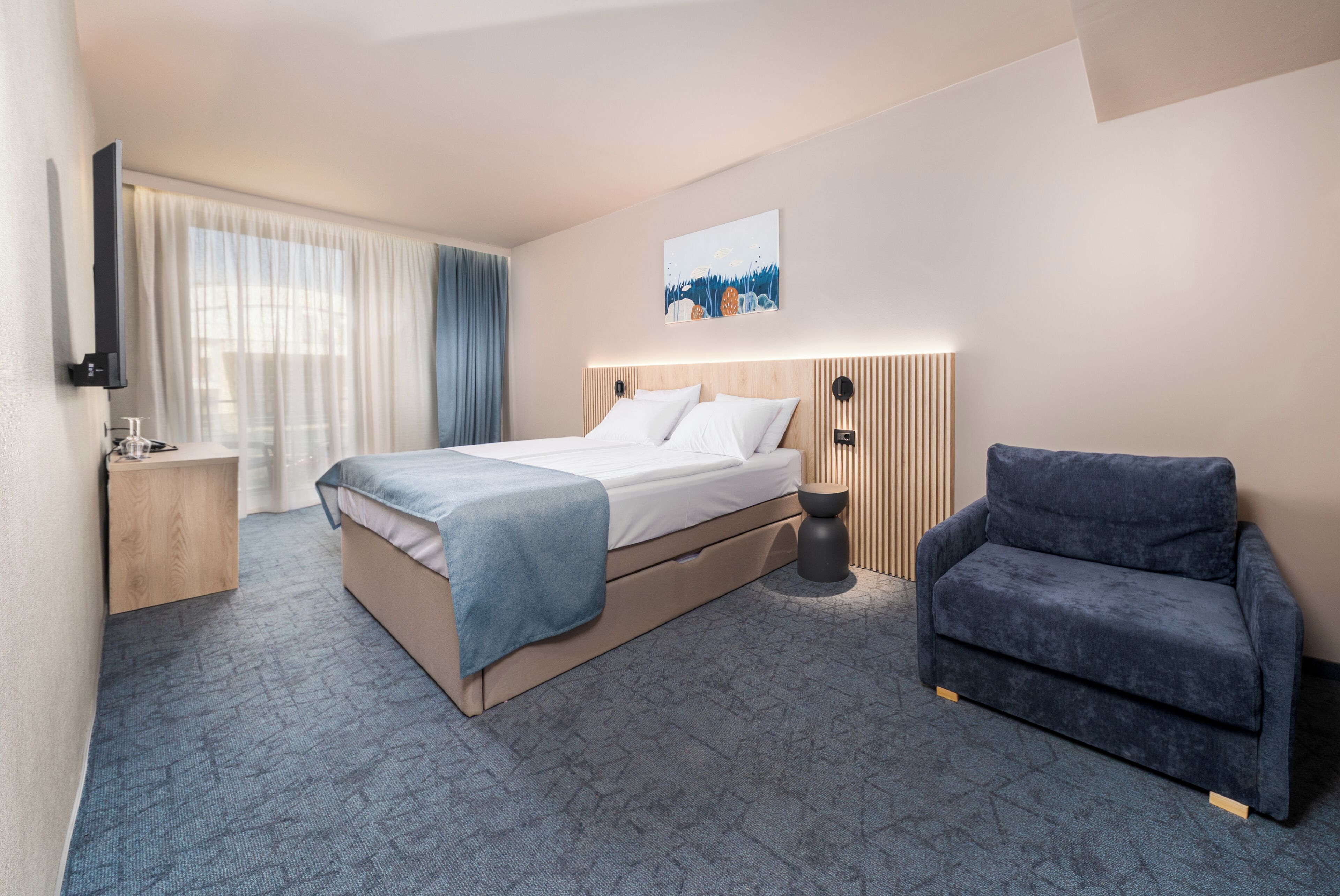 A hotel room with a bed, a chair, a bedside table, a TV, and a painting on the wall.