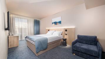 A hotel room with a bed, a chair, a bedside table, a TV, and a painting on the wall.