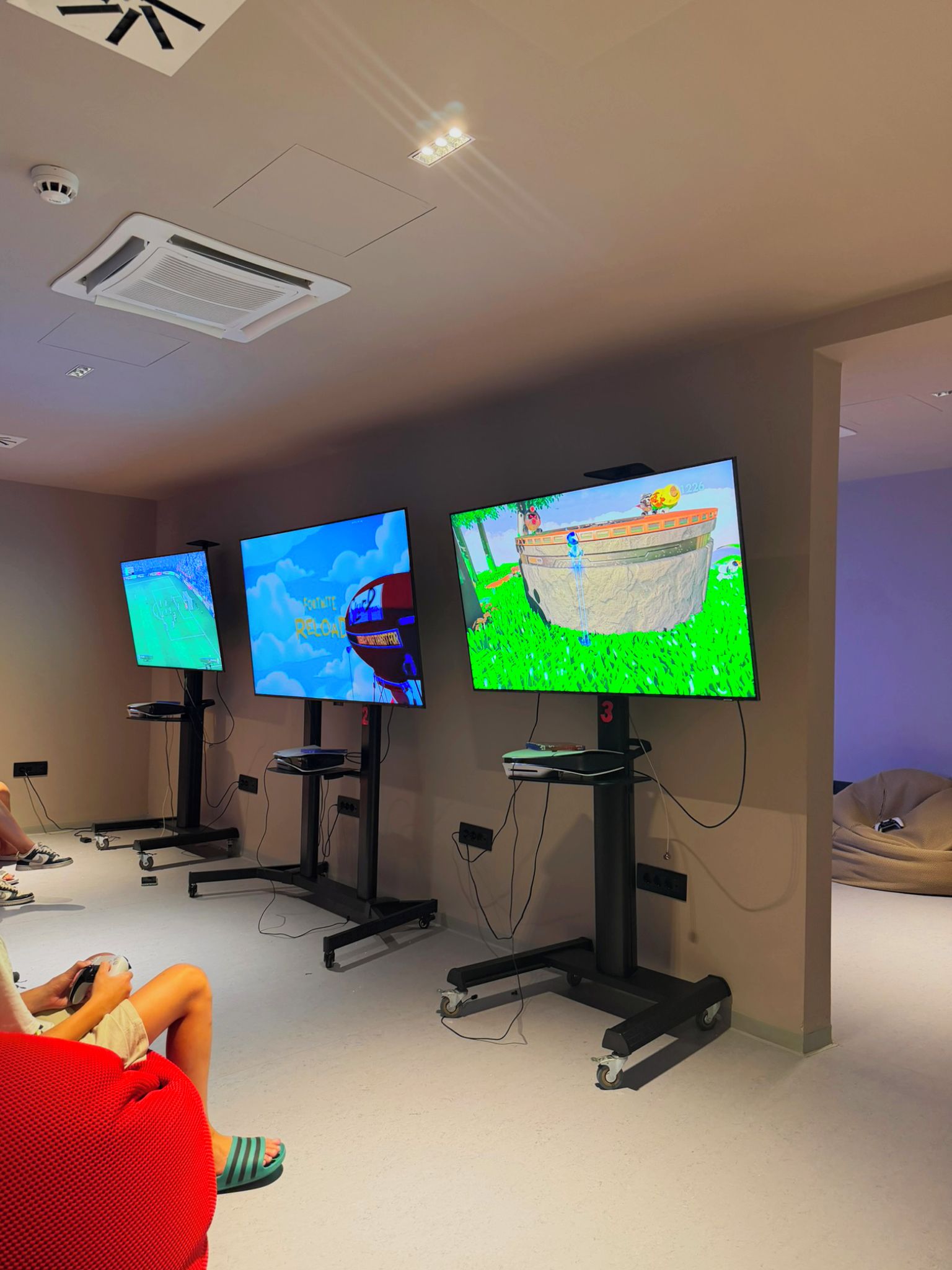 A gaming room with three large flat-screen TVs mounted on stands, a treadmill, and a person sitting on a bean bag.