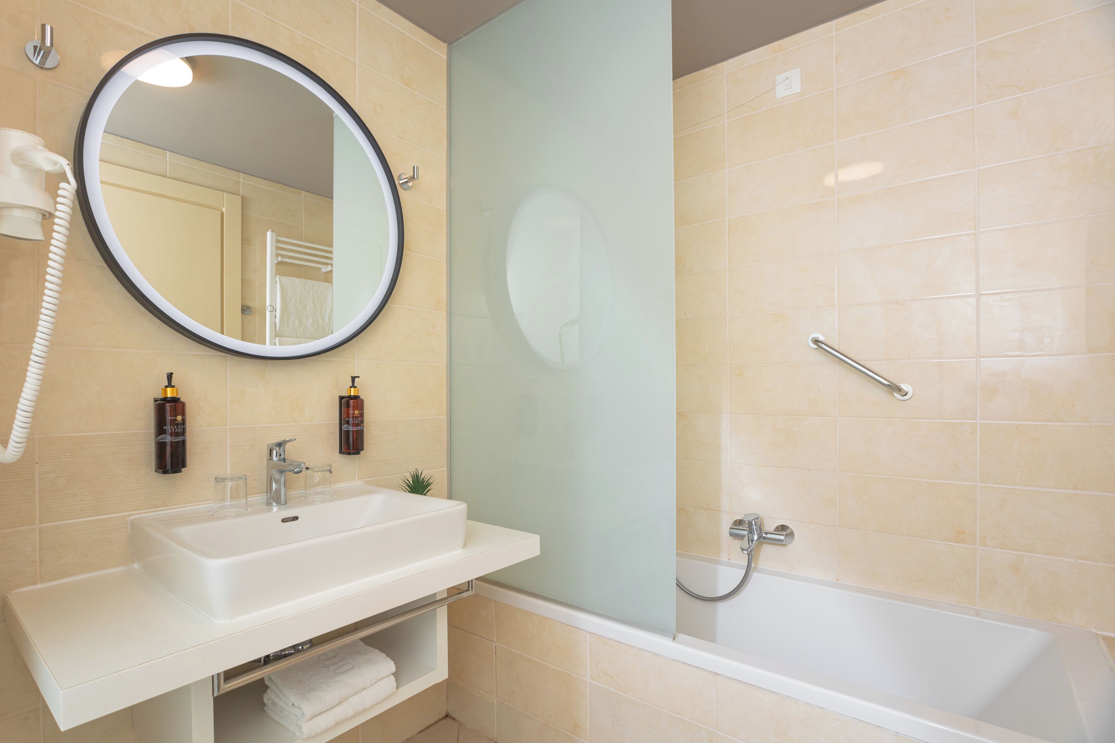 Un baño con lavamanos blanco, espejo redondo, ducha con cabezal de mano y mampara de cristal.