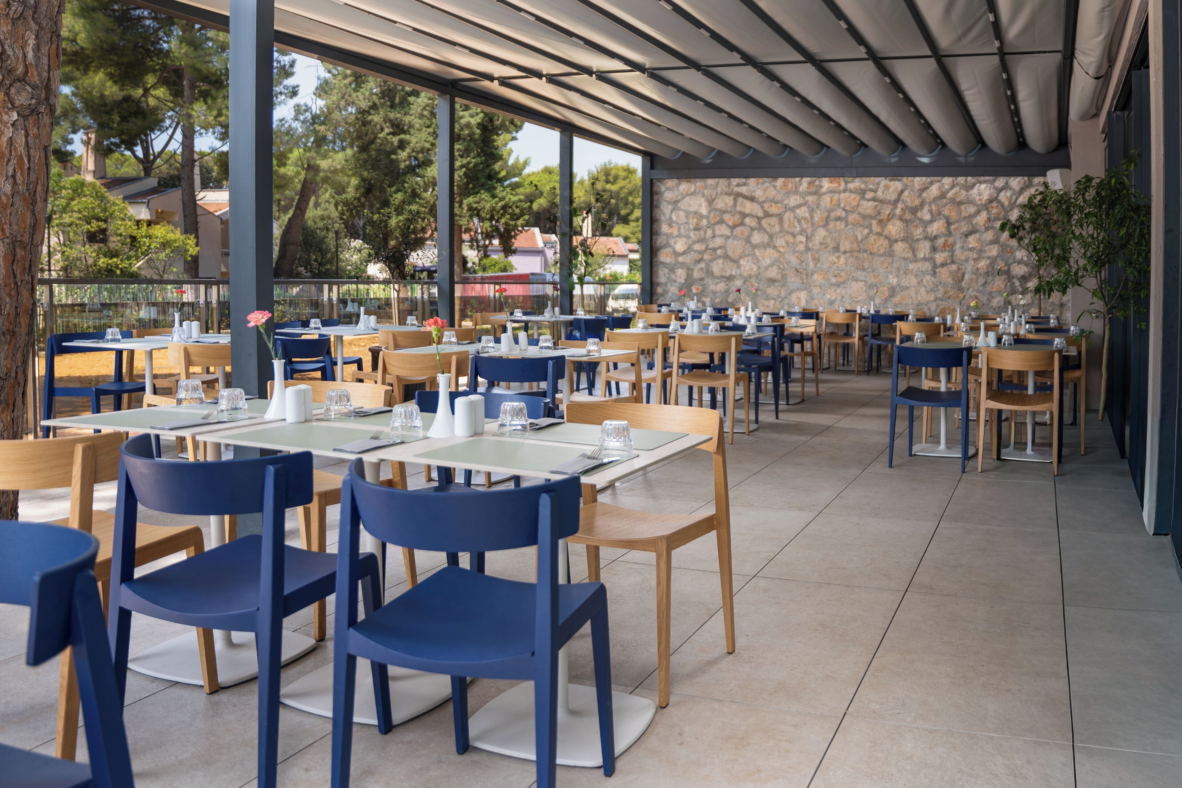 An outdoor dining area with multiple tables and chairs, a stone wall, and a view of trees and buildings in the background.