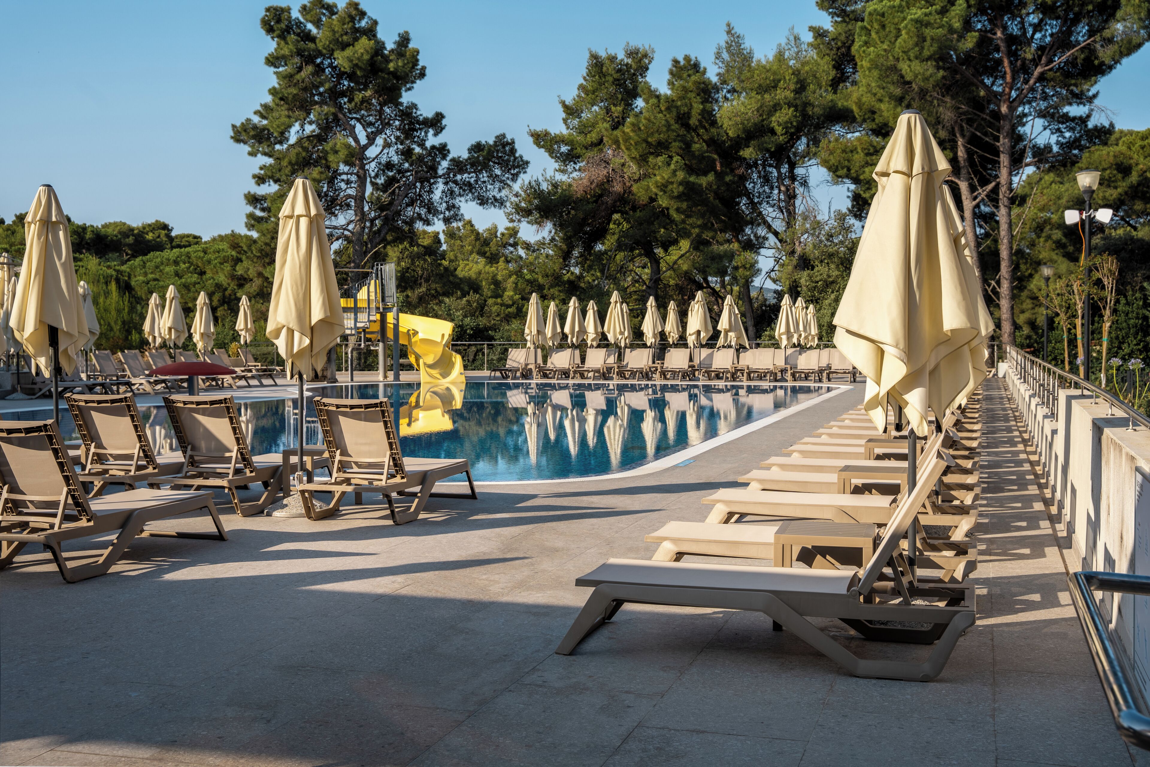A poolside area with sun loungers and umbrellas, surrounded by trees.