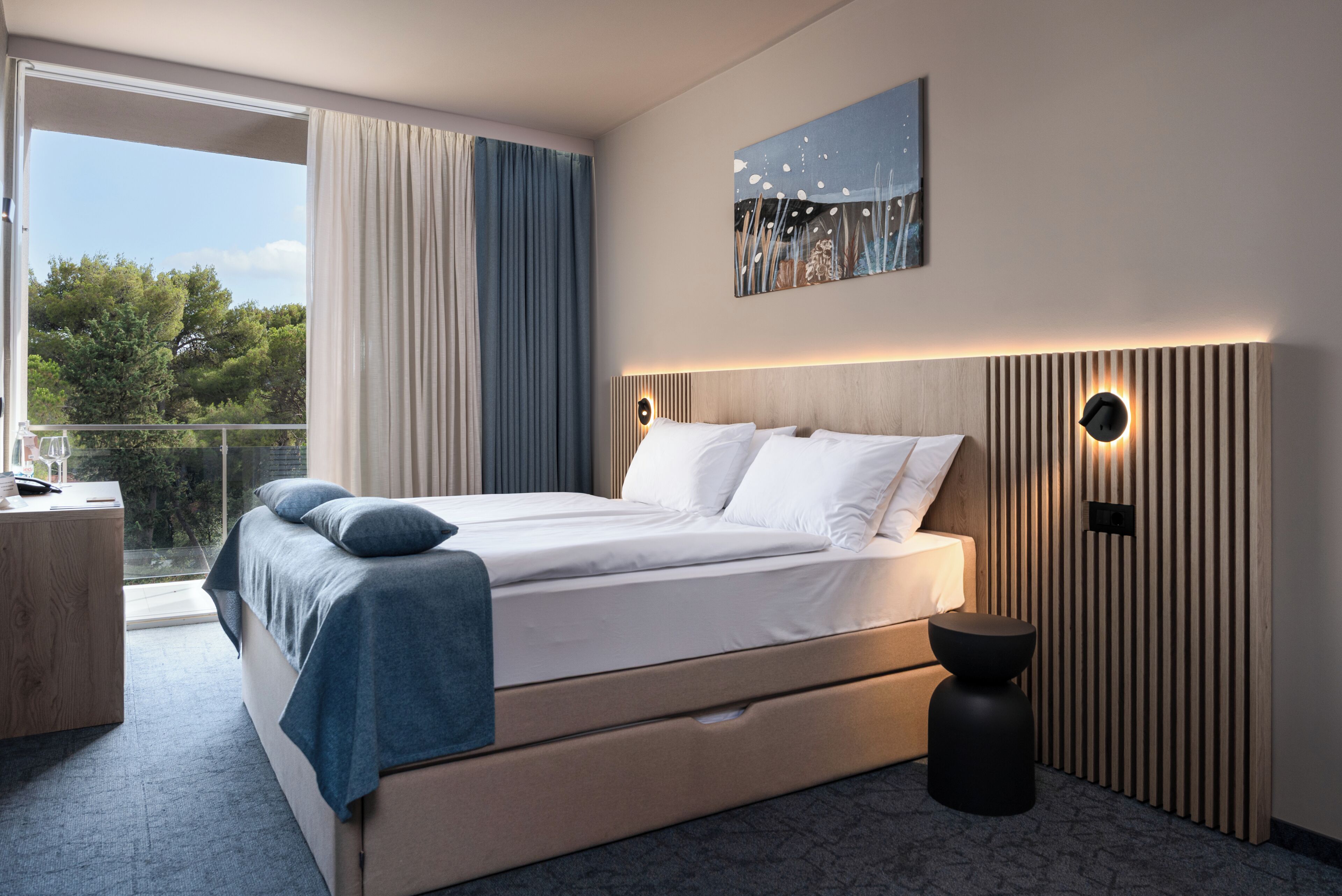 Een moderne slaapkamer met een groot bed, een bijzettafeltje, een klein krukje en uitzicht op het omliggende landschap.