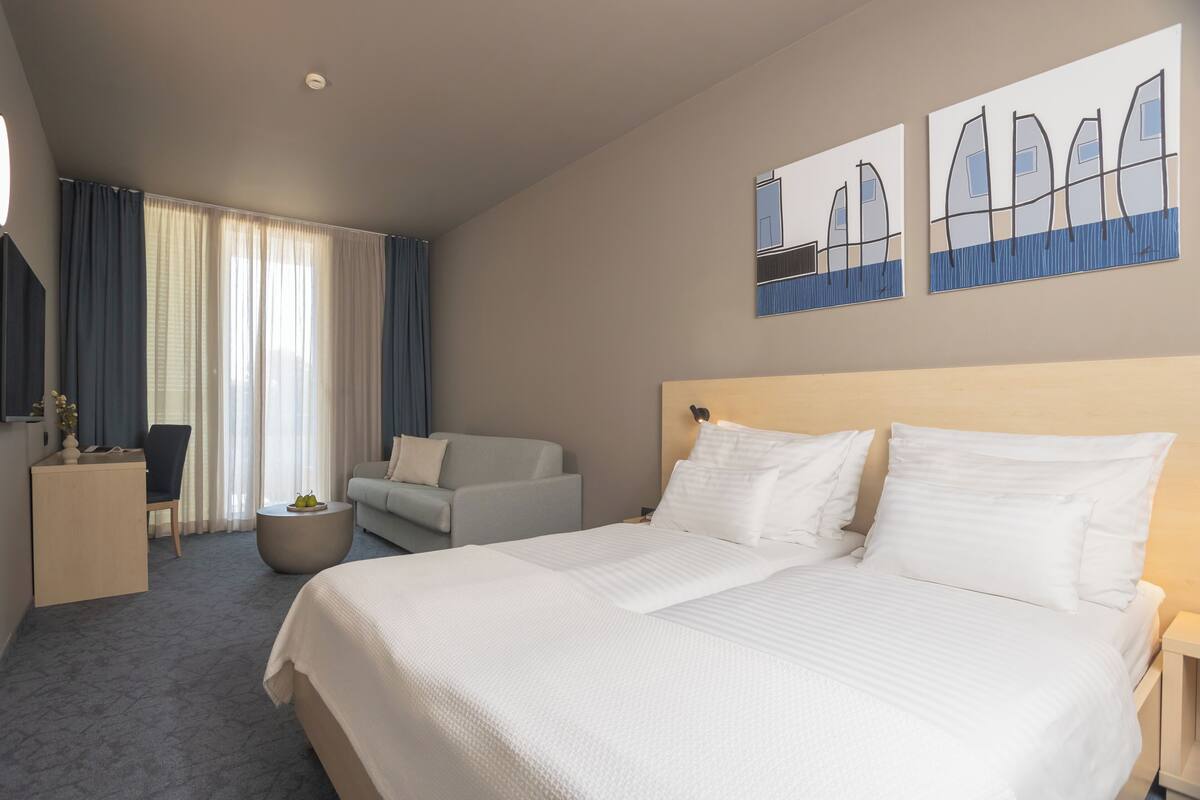 A hotel room with a large bed, a sofa, a round coffee table, and a desk. There are two paintings on the wall and a window with curtains.