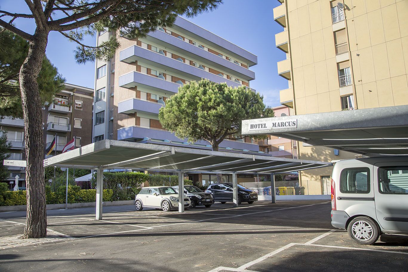 A hotel parking area with a covered car park, a tree, and a multi-story building in the background.