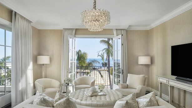 Een ruime woonkamer met een grote, ronde, knopensofa, twee fauteuils, een televisie en een balkon met uitzicht op zee.