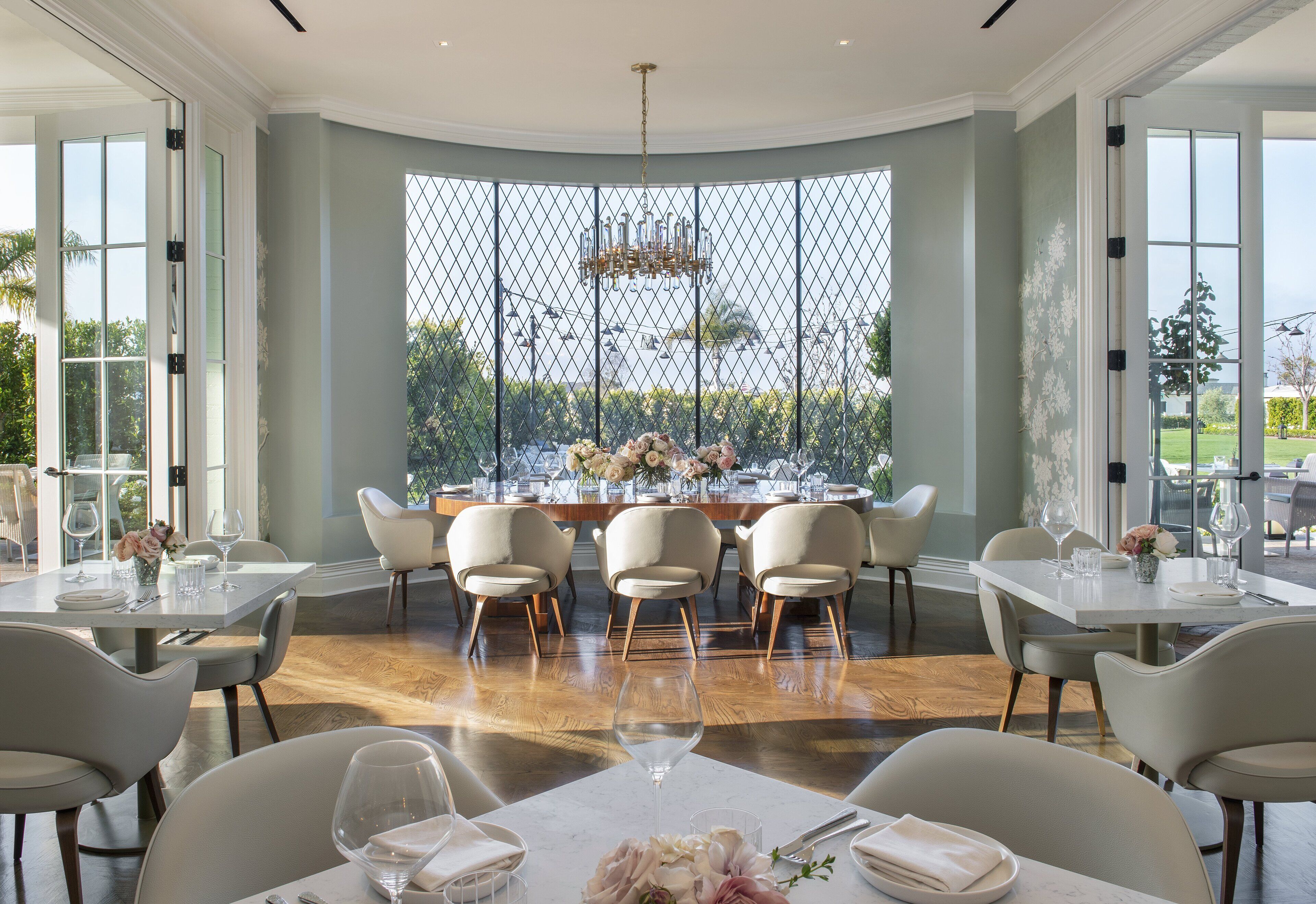 A dining area with a large table set for six, surrounded by six chairs, with a view through a patterned window.