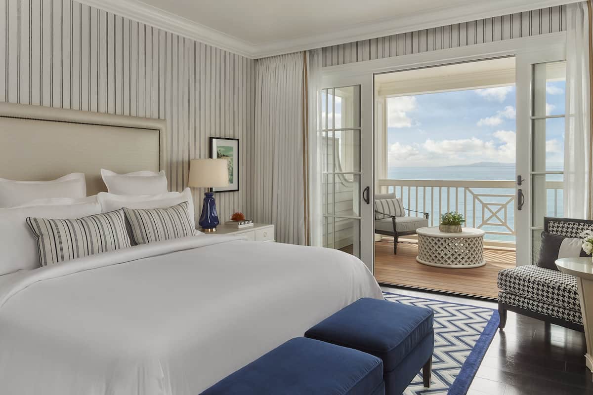 Een hotelkamer met een groot bed, een balkon met uitzicht op zee, een klein rond tafeltje en een stoel.