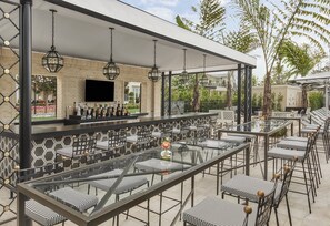 An outdoor bar area with hexagonal patterned walls, a long counter with bottles, and multiple tables with striped cushions.