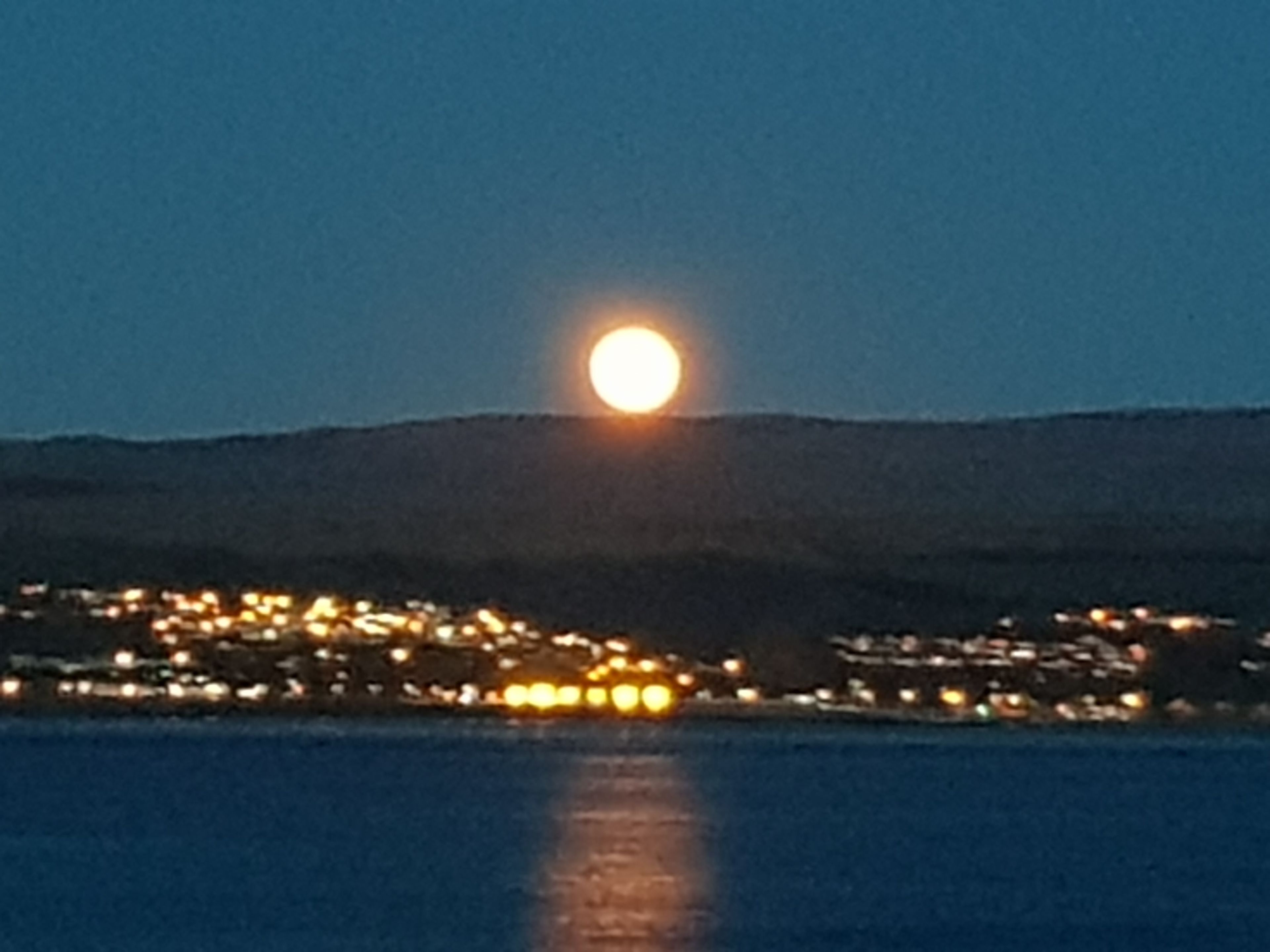 Un pueblo costero de noche, con luna visible y un cuerpo de agua que refleja las luces de la ciudad.