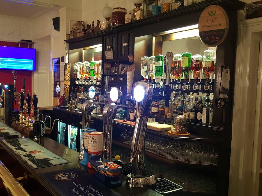 Zona de bar con múltiples grifos de cerveza, una variedad de botellas de licores y una pantalla de televisión mostrando contenido.