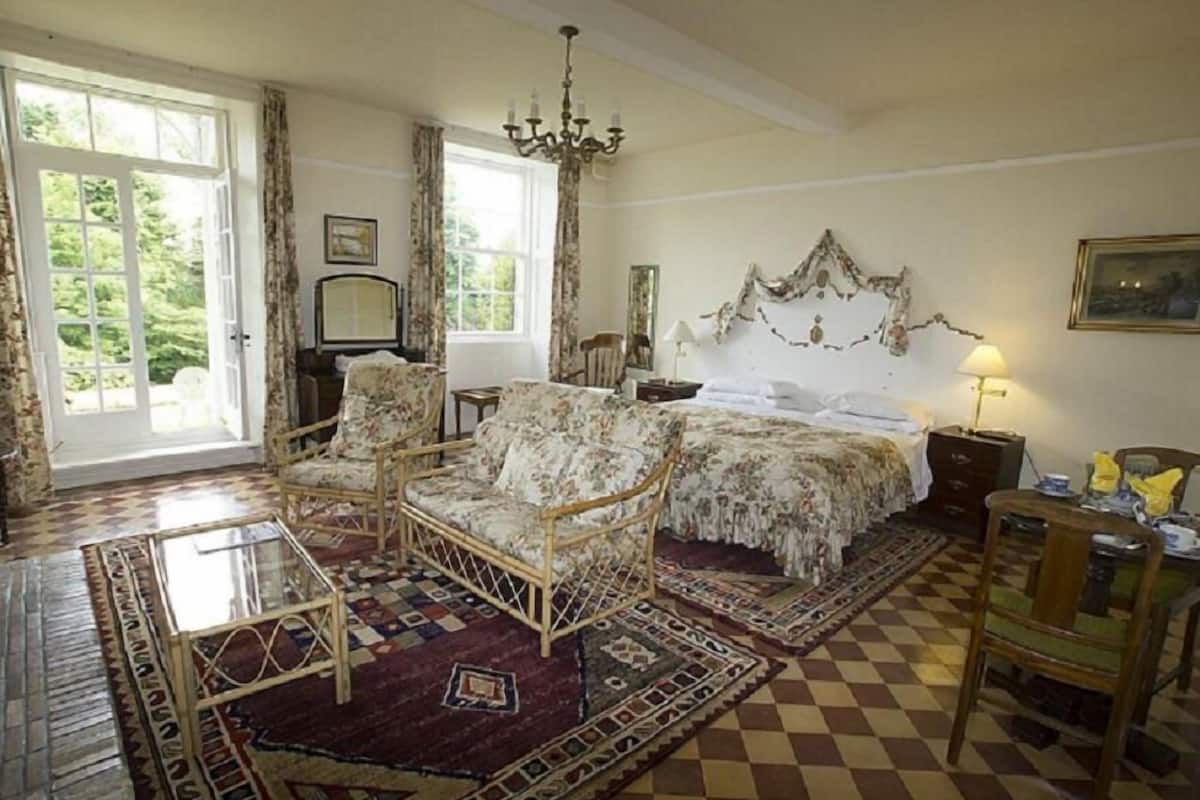 A room with a checkered floor, a bed with floral bedding, a wooden chair, a glass table, and a chandelier.