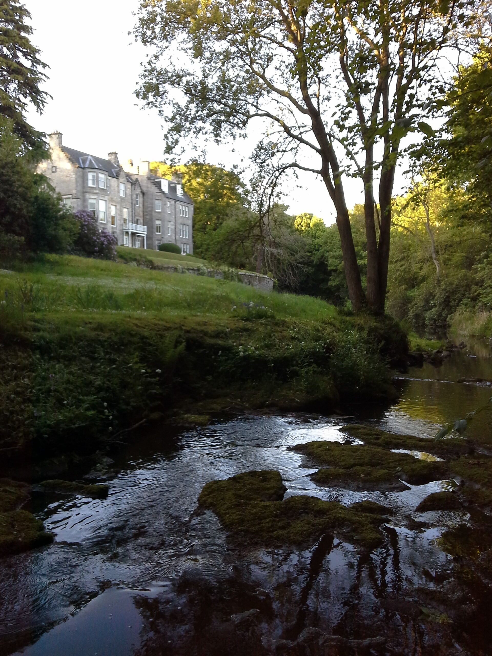 A river with moss-covered rocks, a grassy bank, and a stone building with multiple windows in the background.