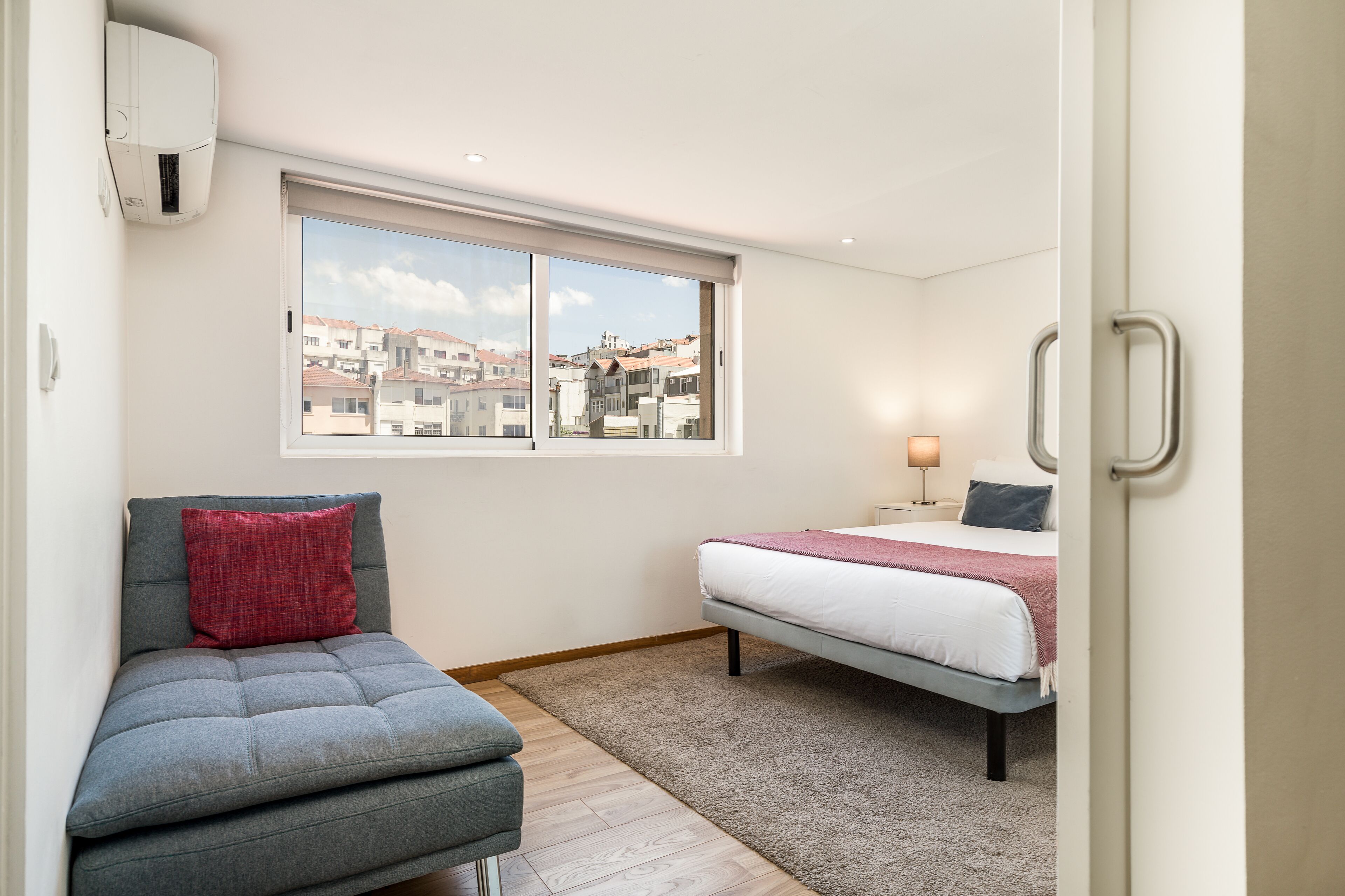 A bedroom with a bed, a sofa, a window with a city view, and an air conditioning unit.