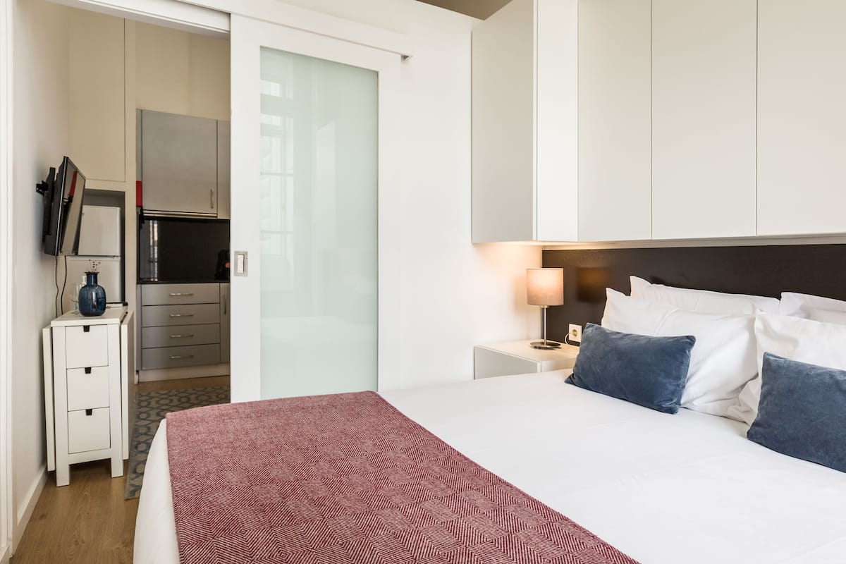 A hotel room with a bed, bedside table, lamp, and a view of a kitchen area.