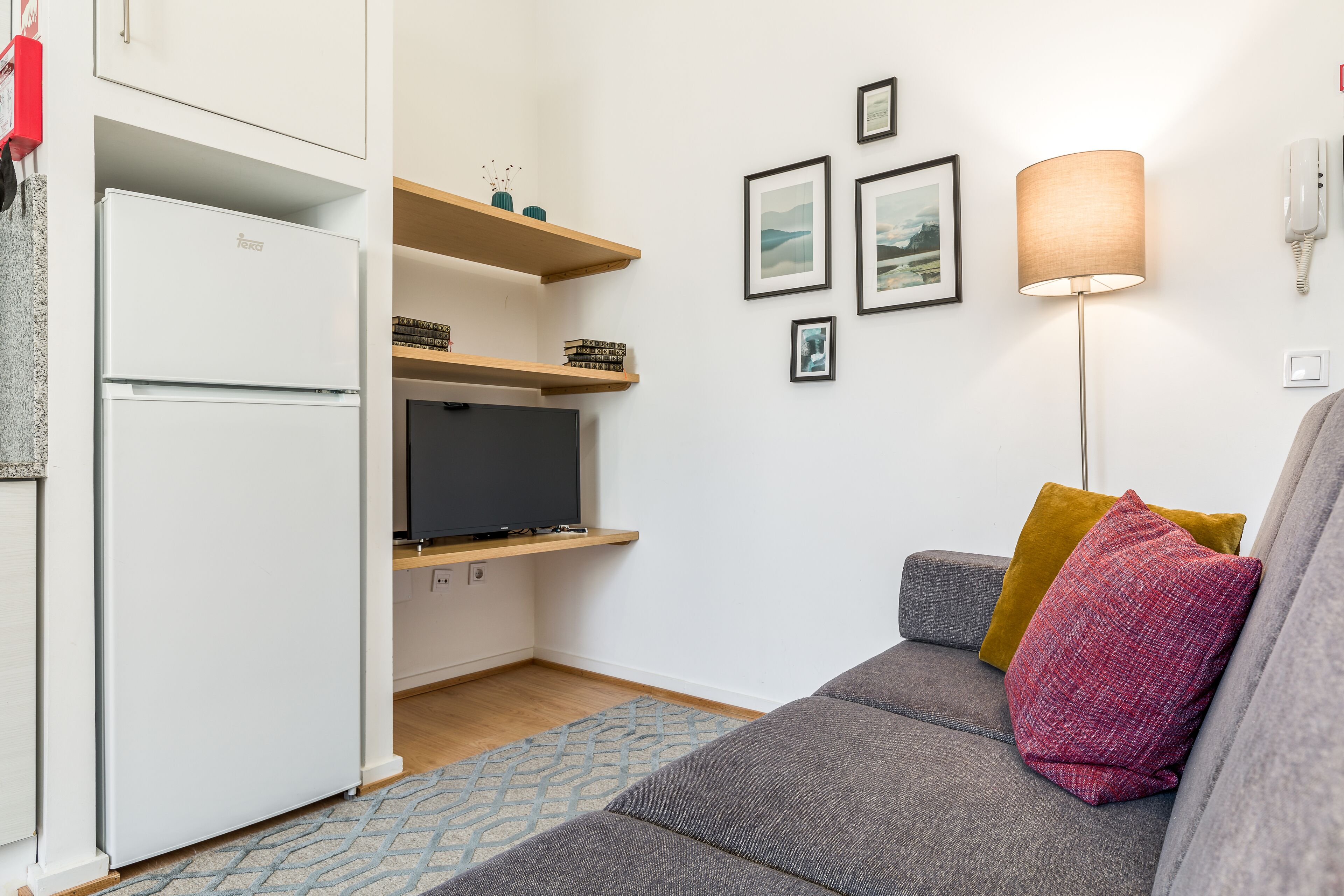 Un espacio habitable compacto con kitchenette, un sofá gris con cojines y un televisor montado en la pared.