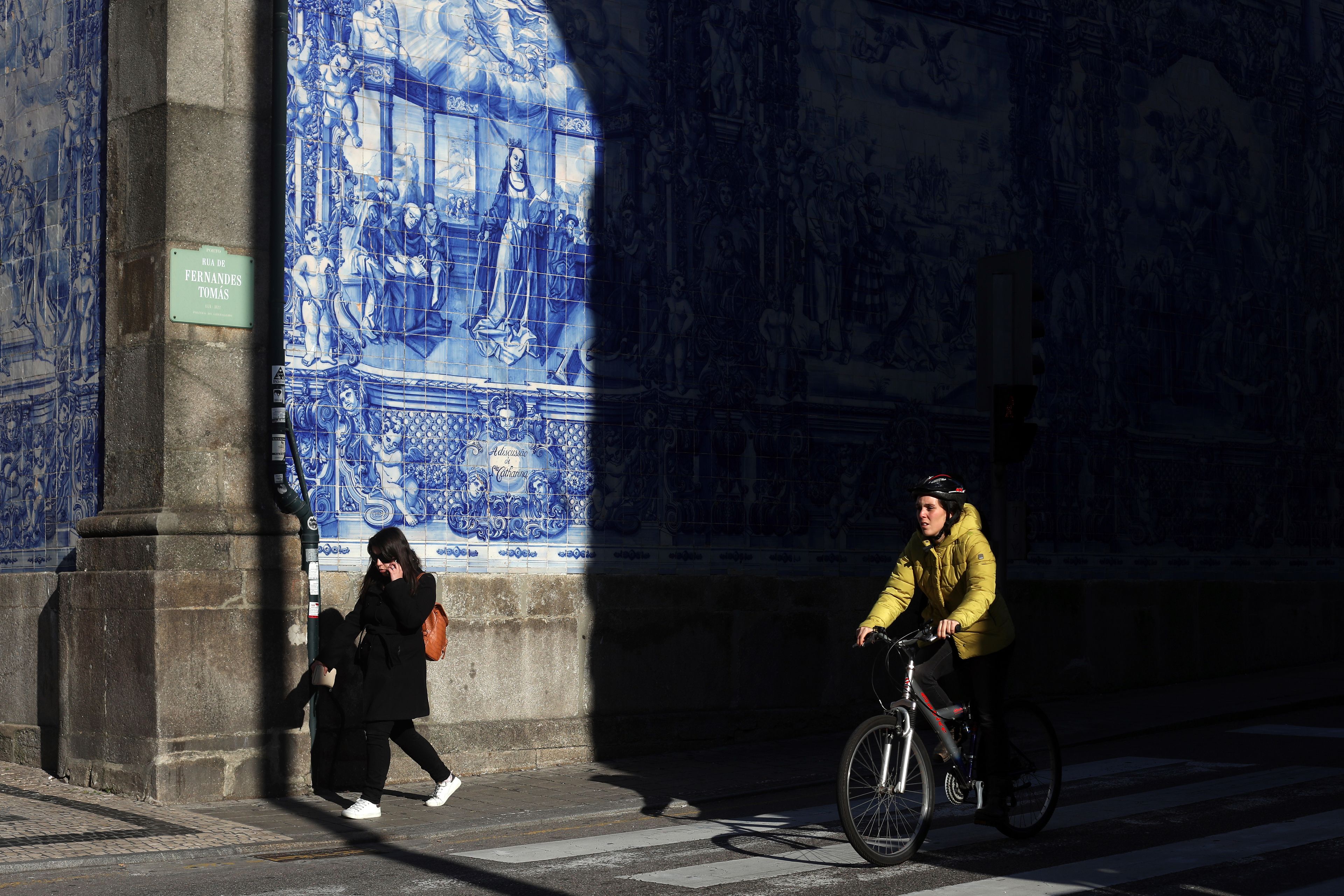A street scene with a tiled wall featuring intricate blue designs, a sign reading "Rua de Fernandas Tomas," and a cyclist in a yellow jacket.