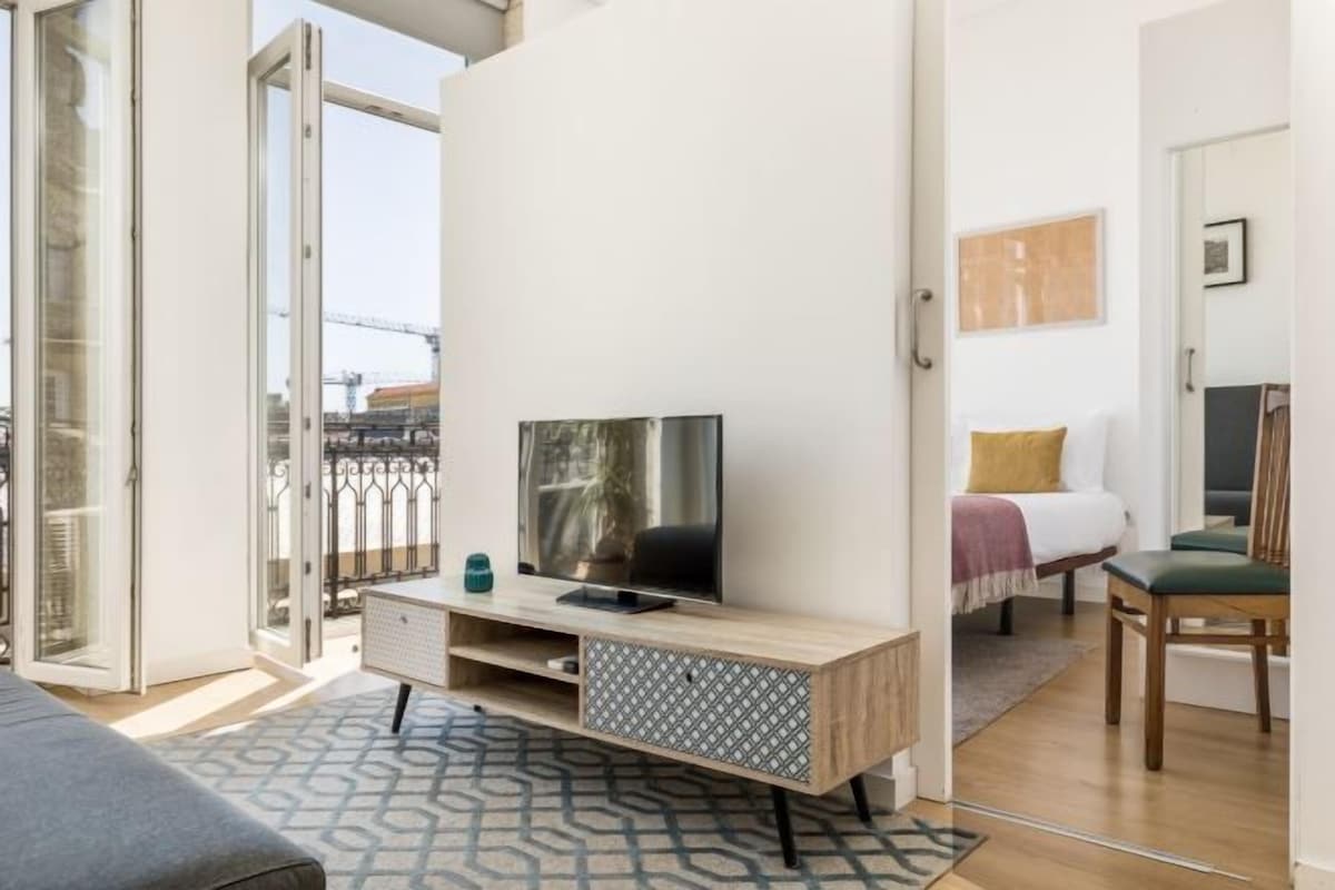 Una habitación de hotel moderna con un ventanal grande, un televisor de pantalla plana sobre un mueble de madera, una cama con una silla verde y un balcón con vistas a la ciudad.