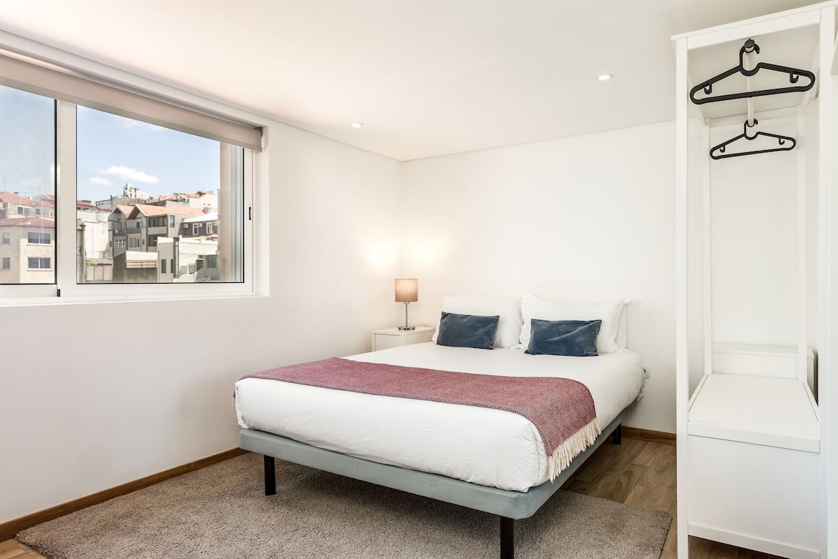 A bedroom with a large window, a bed with white bedding, a red throw blanket, a nightstand with a lamp, and a wardrobe with hangers.