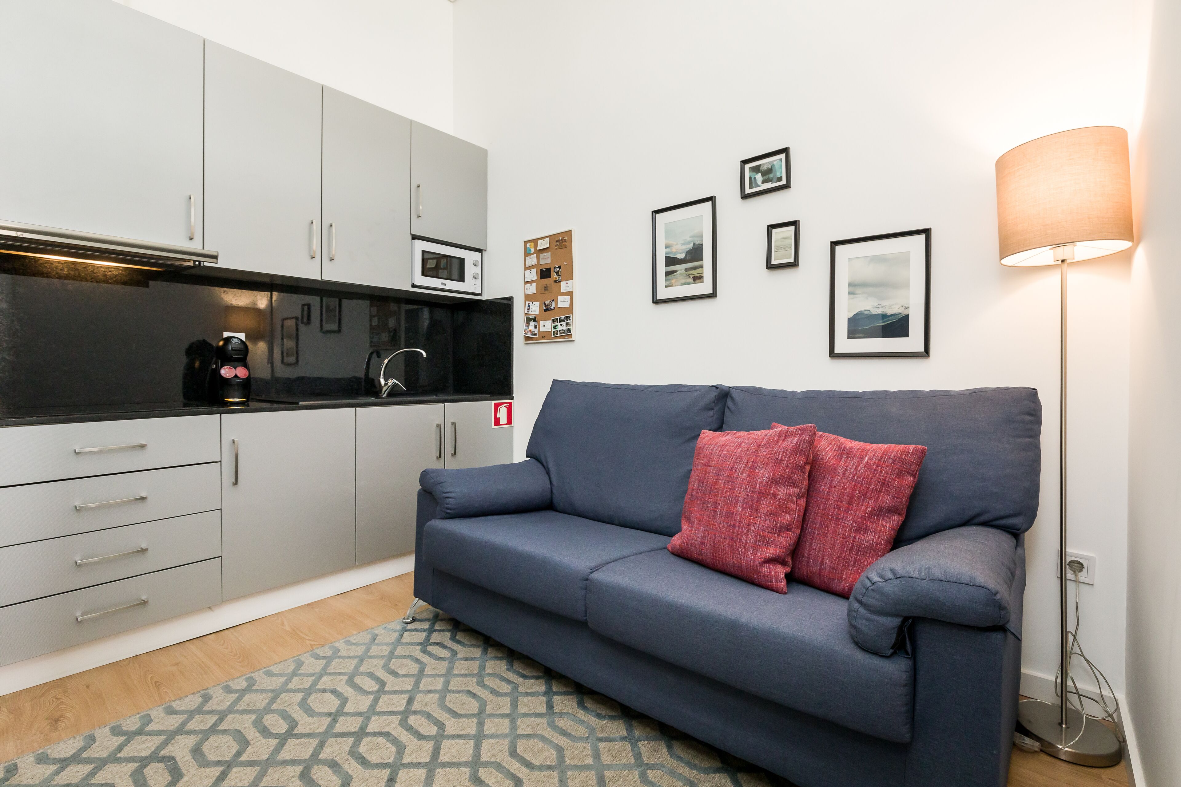 A modern kitchen with grey cabinets, a black countertop, and a microwave. There is a blue sofa with a red pillow and a patterned rug.