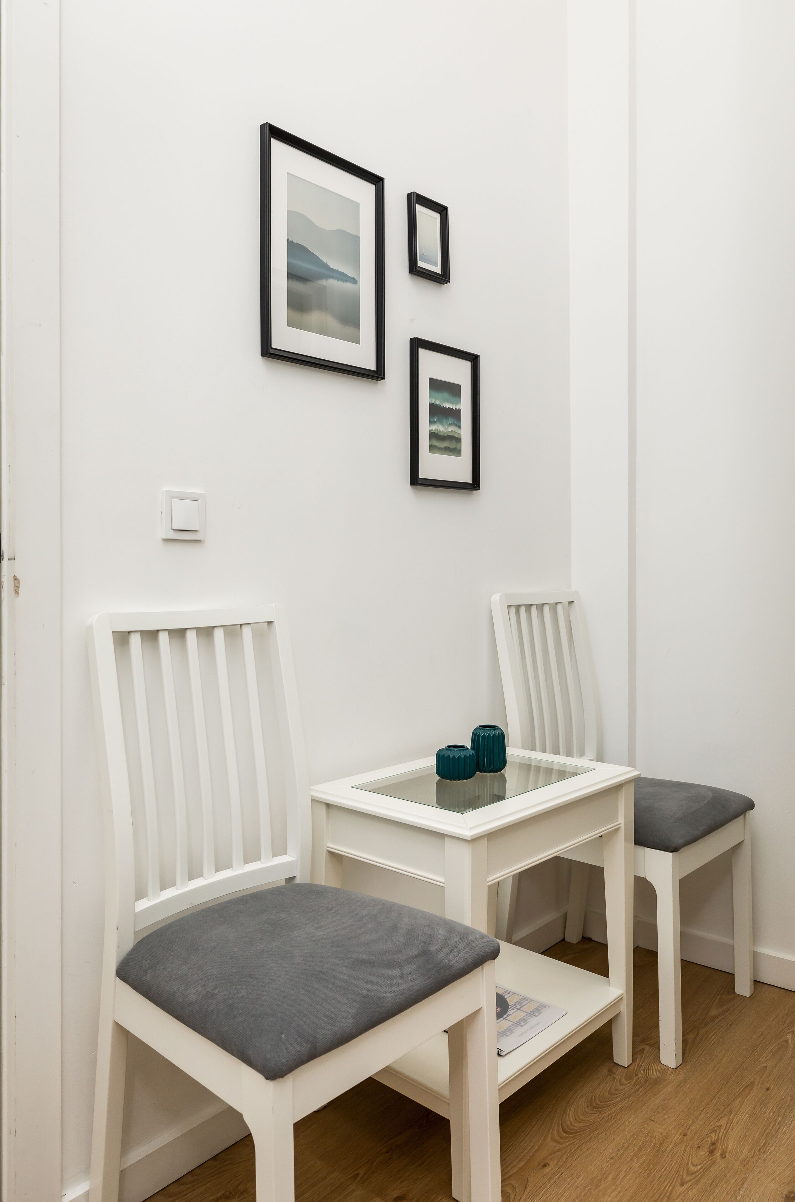 A room with two white chairs with grey cushions, a small white table, and two framed pictures on the wall.