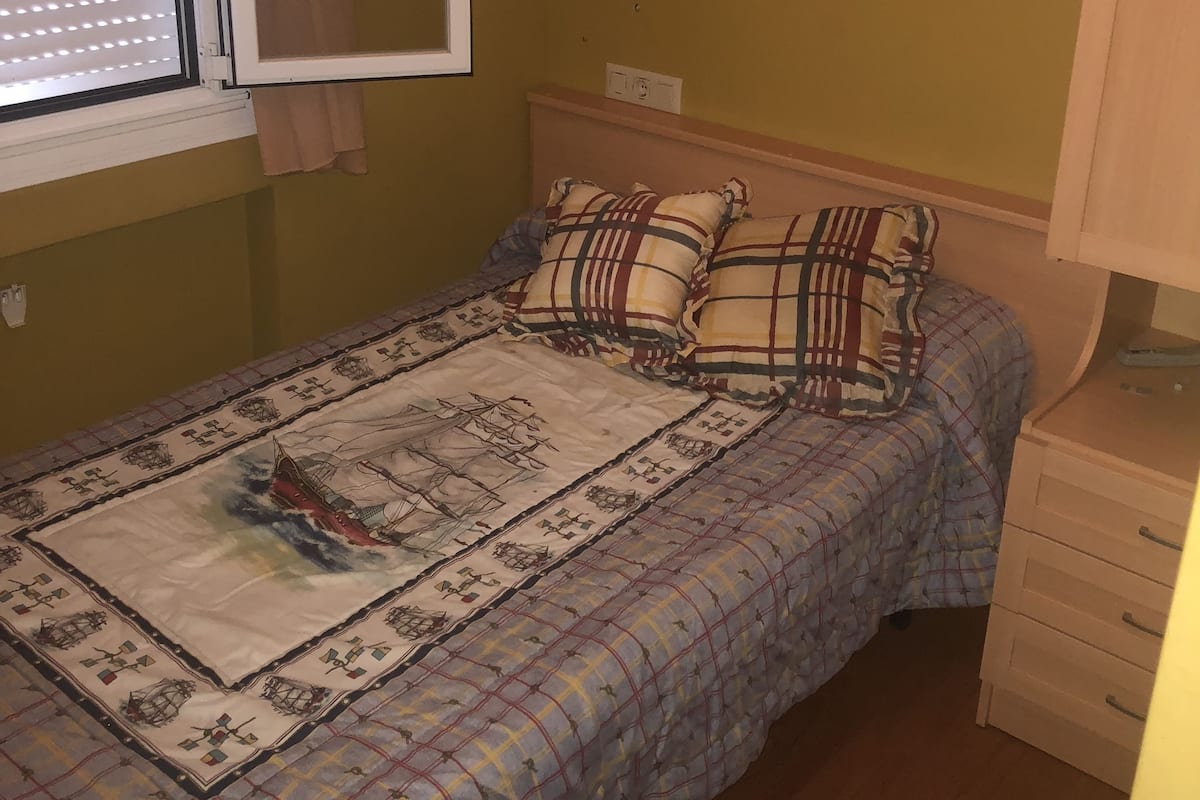 A bedroom with a bed, a nightstand, a window with blinds, and a wall socket.