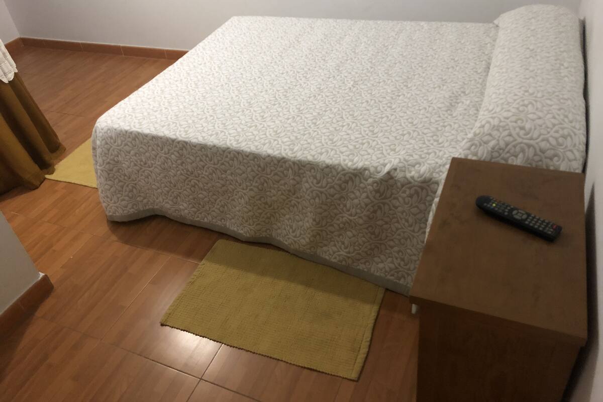 A neatly made bed with a patterned bedspread, a brown bedside table with a remote control, and a yellow rug on a wooden floor.