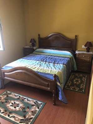 Quarto com cama de madeira, duas mesas de cabeceira, luminária, tapete estampado e janela.