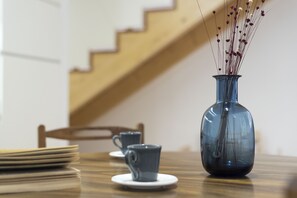 A dining table with a blue vase containing dried flowers, a stack of plates, and two blue cups on saucers.