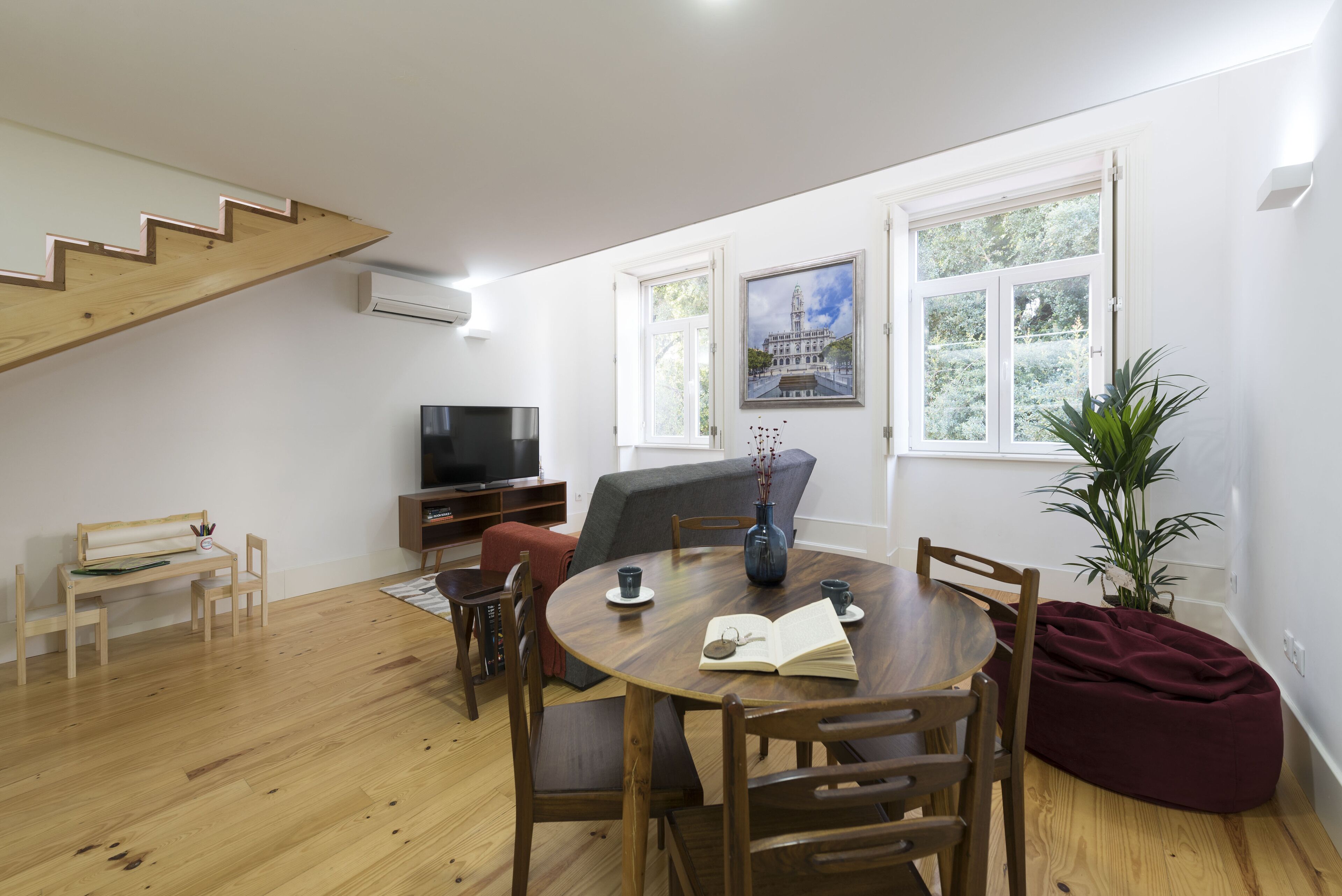 Uma sala de estar moderna com escada de madeira, mesa de jantar com cadeiras, sofá, televisão e uma janela grande com vista para vegetação.