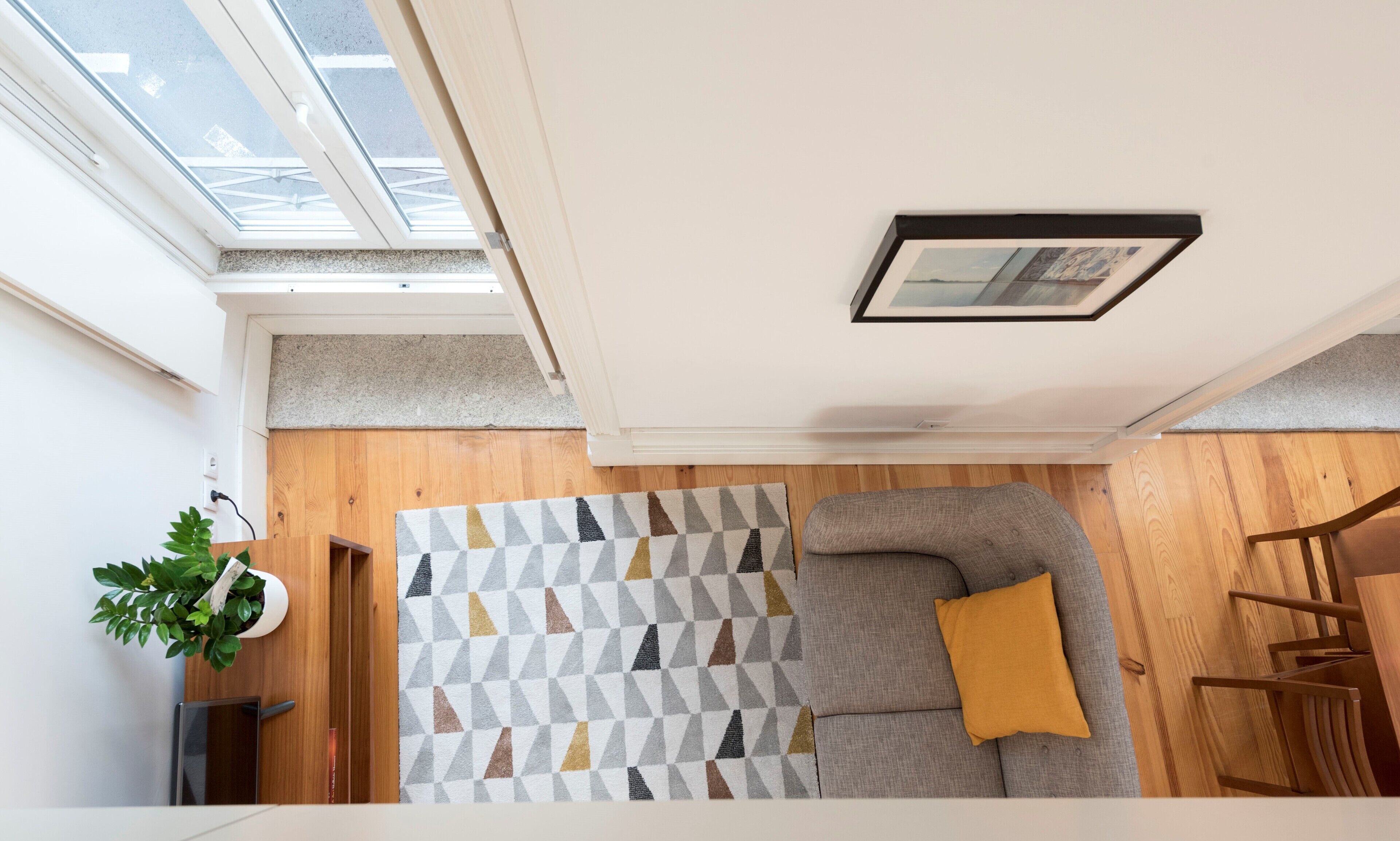 A modern living room with a geometric-patterned rug, a grey sofa with a mustard pillow, a wooden floor, and a skylight.
