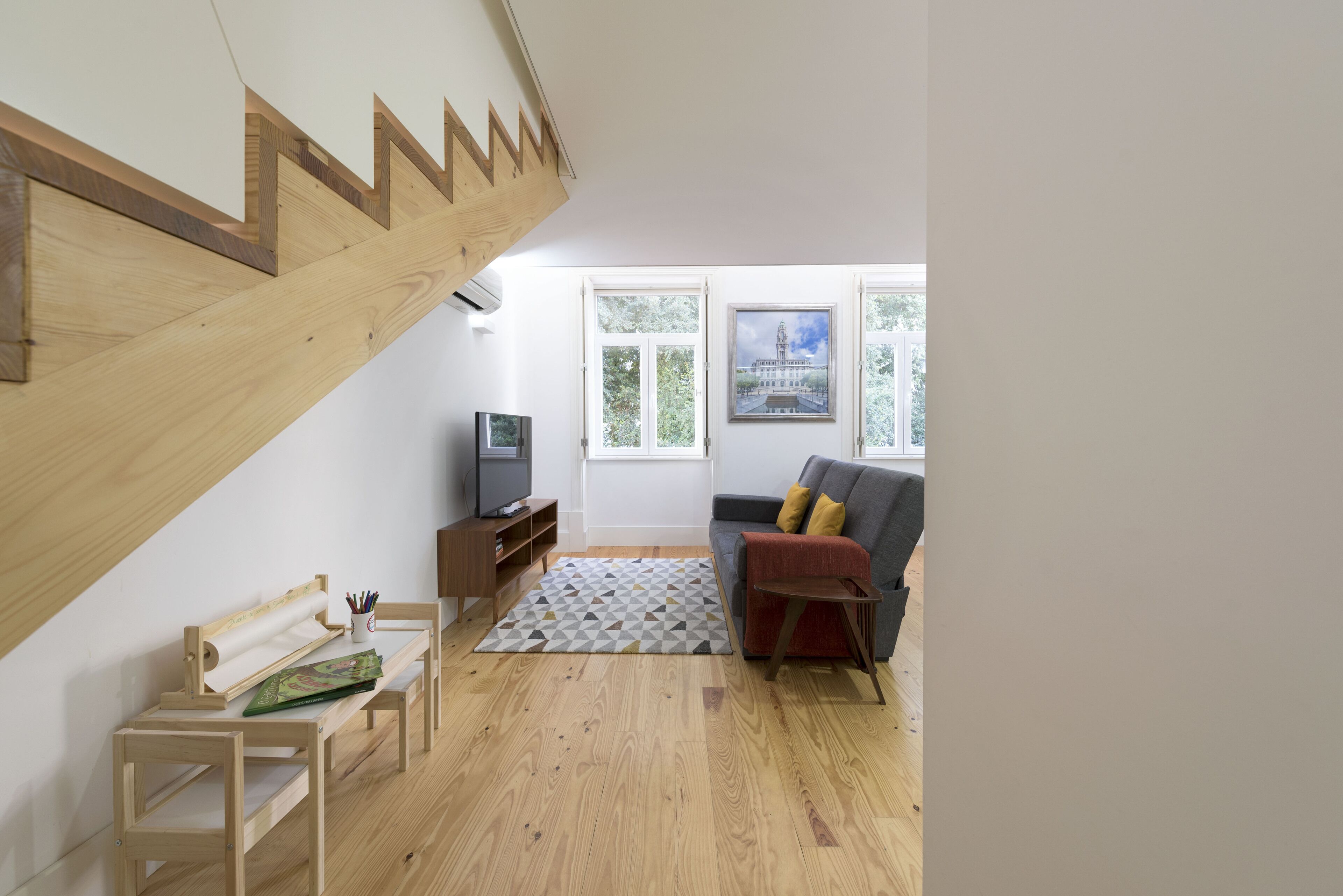 A modern living room with a wooden staircase, a grey sofa with yellow cushions, a television, and a patterned rug.